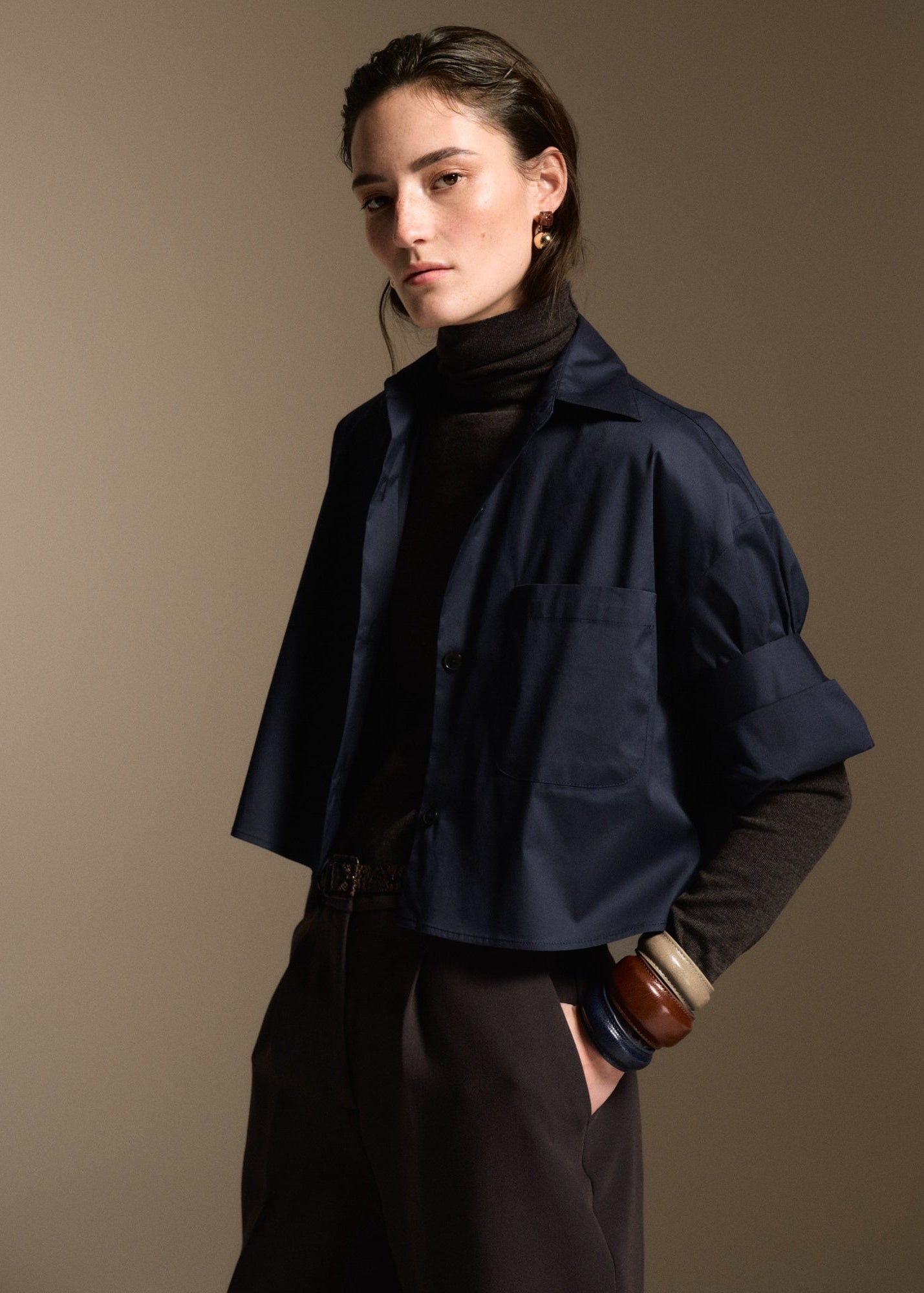 A model wearing a cropped navy blue button-up shirt jacket over a black turtleneck, paired with dark trousers, set against a solid warm brown studio background.