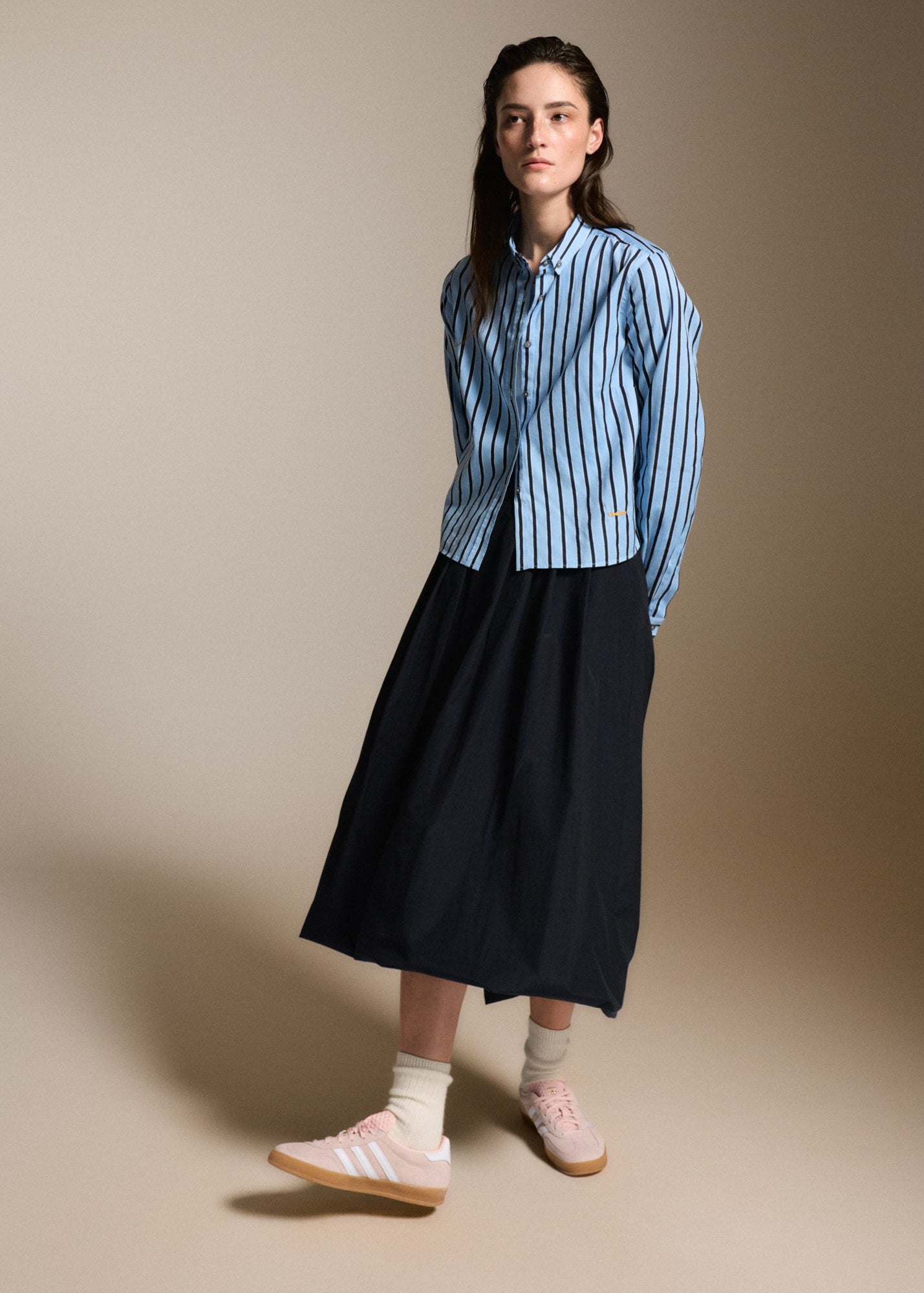 A model stands against a warm beige studio background wearing a light blue and white vertically striped button-down shirt, a dark navy midi skirt, white socks, and light pink sneakers.