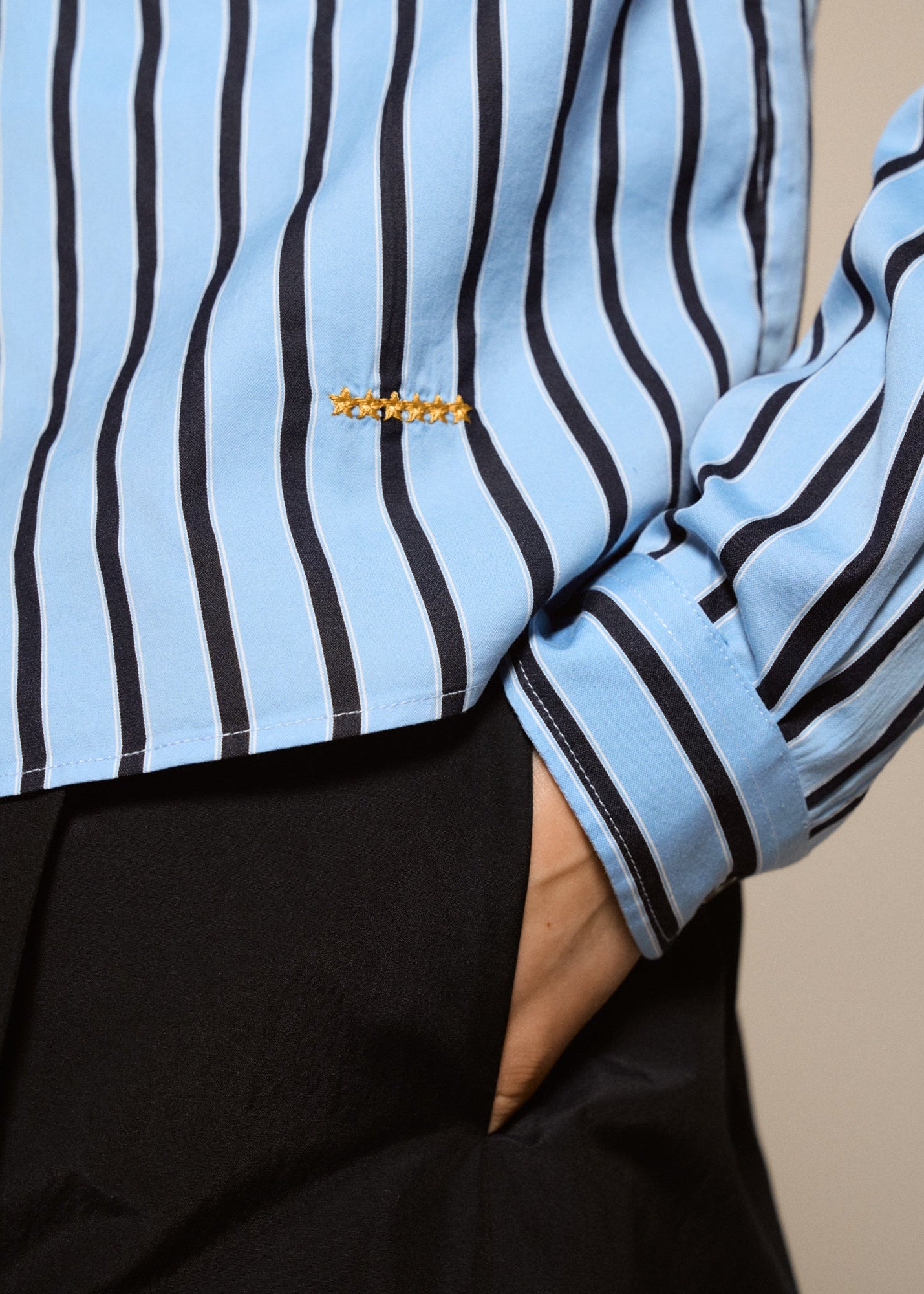 Close-up of a person wearing a light blue and black vertically striped long-sleeve shirt with a small gold embroidered detail on the chest, paired with black trousers.
