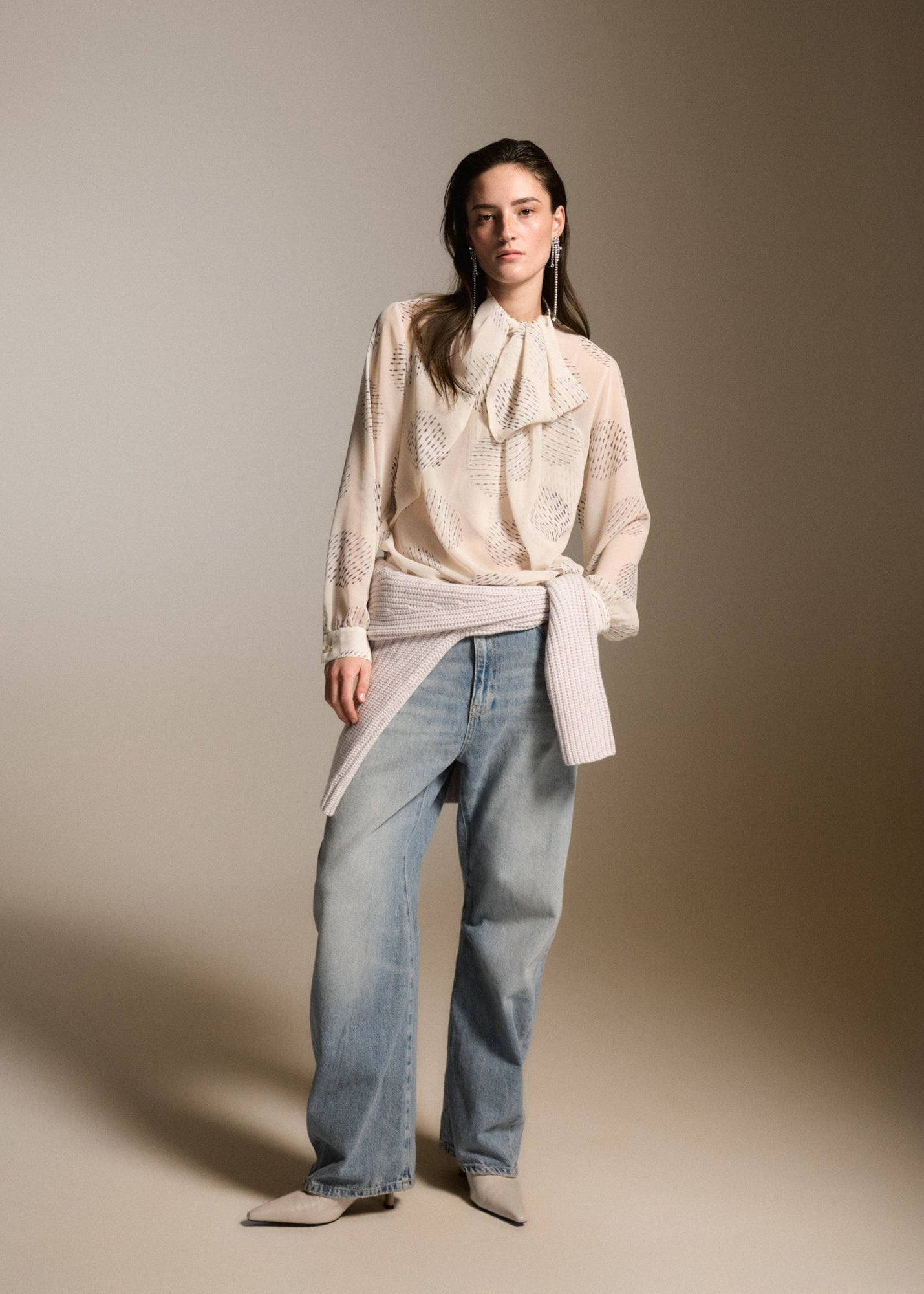 A model stands against a warm beige studio backdrop wearing a cream-colored lace blouse with a large bow at the collar, tied with a ribbed knit belt, paired with wide-leg light blue jeans and pointed beige shoes.
