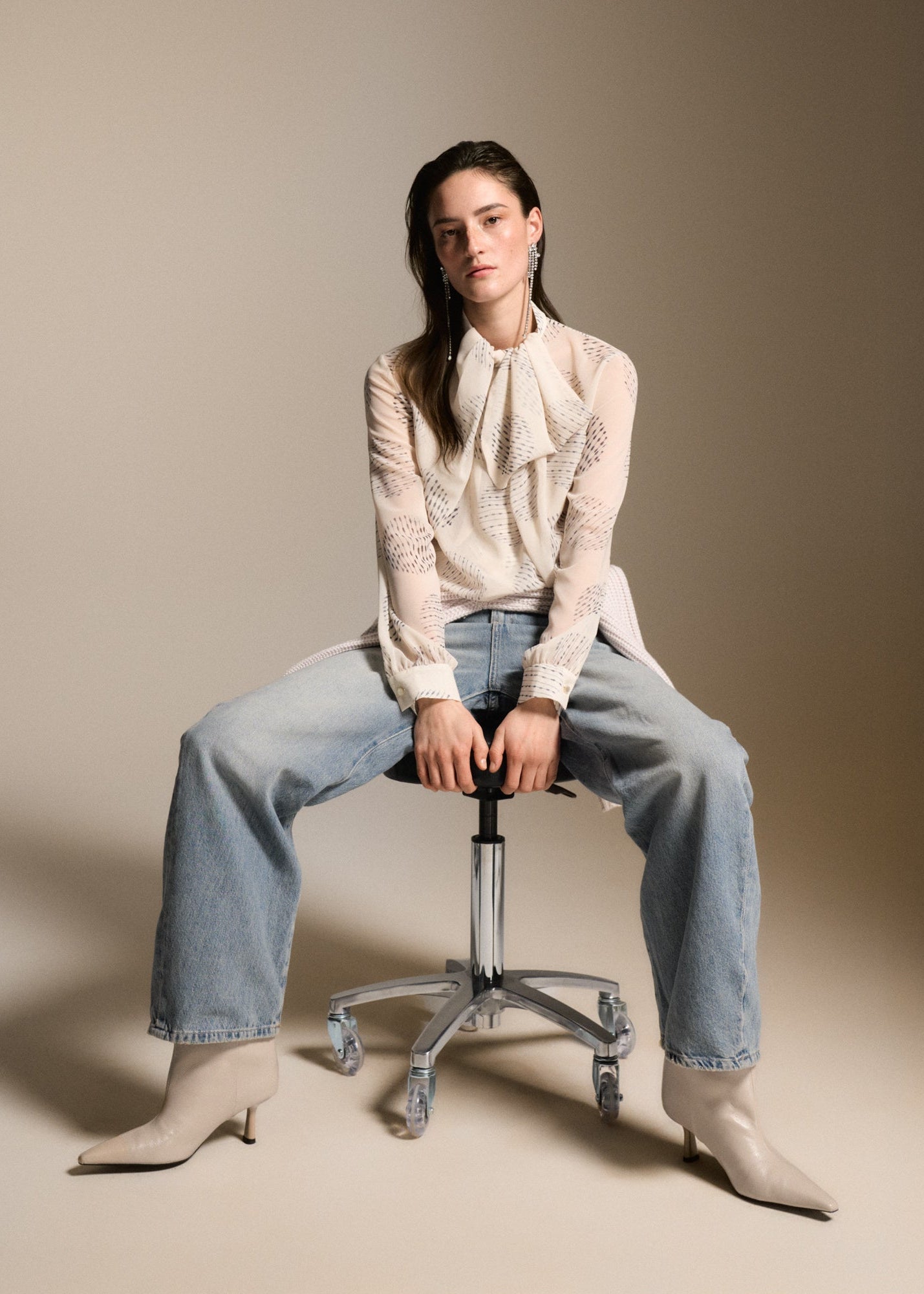 A model sits on a chrome rolling stool wearing a cream lace blouse, light-wash wide-leg jeans, and beige pointed-toe heeled boots against a solid beige studio background.