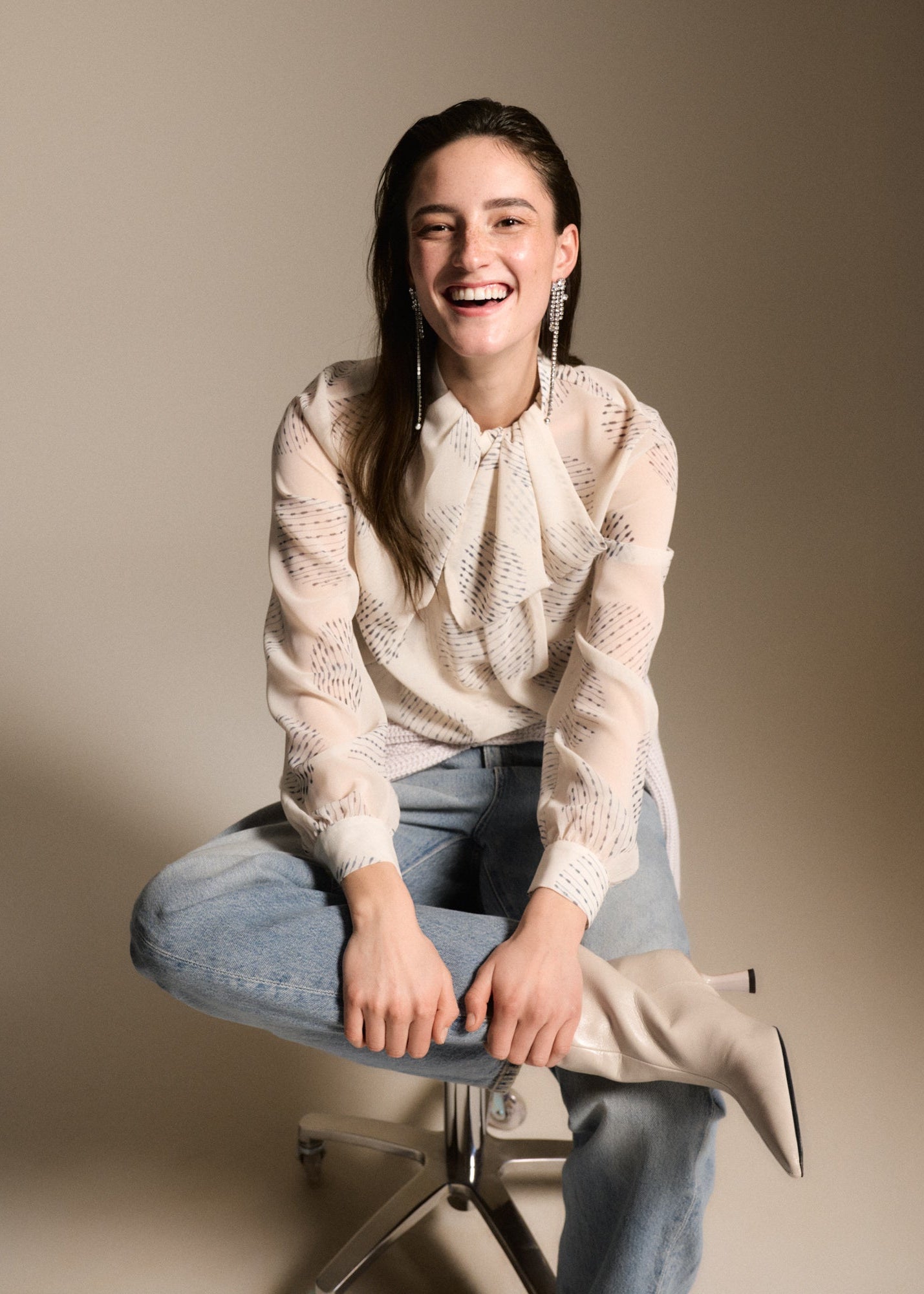 A woman sitting on a metal stool wearing a cream-colored, semi-sheer patterned blouse, light-wash blue jeans, and beige ankle boots against a solid beige studio background.