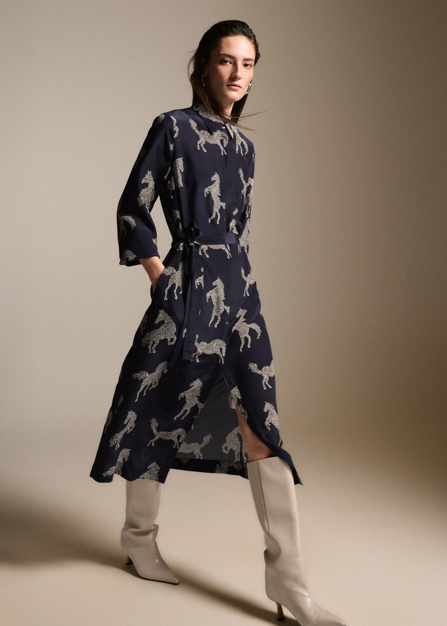 A model wears a navy blue midi dress with a white horse pattern, styled with off-white pointed-toe boots against a neutral beige studio background.