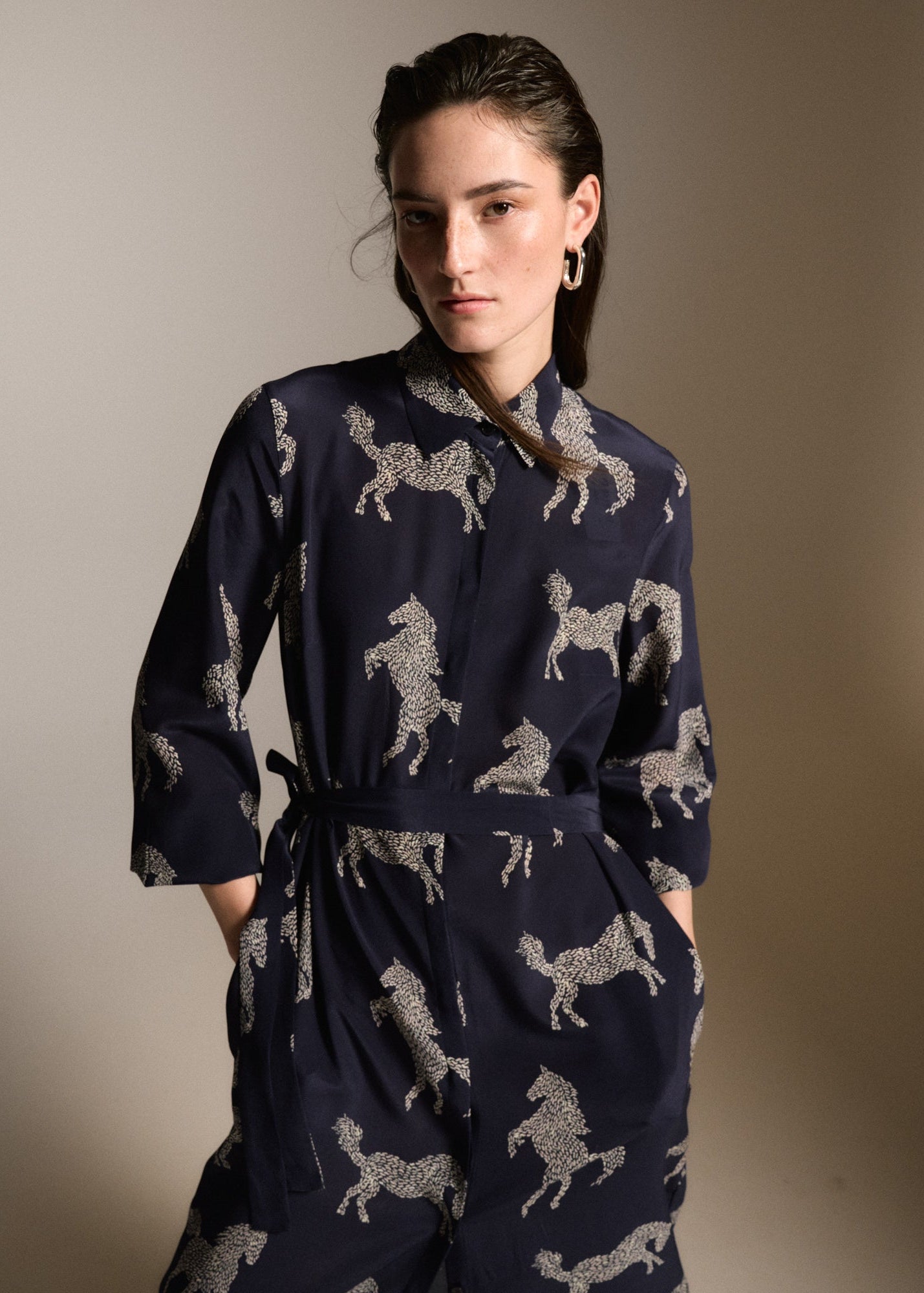 A woman modeling a navy blue wrap-style jumpsuit with a beige horse pattern, photographed against a neutral beige studio background.