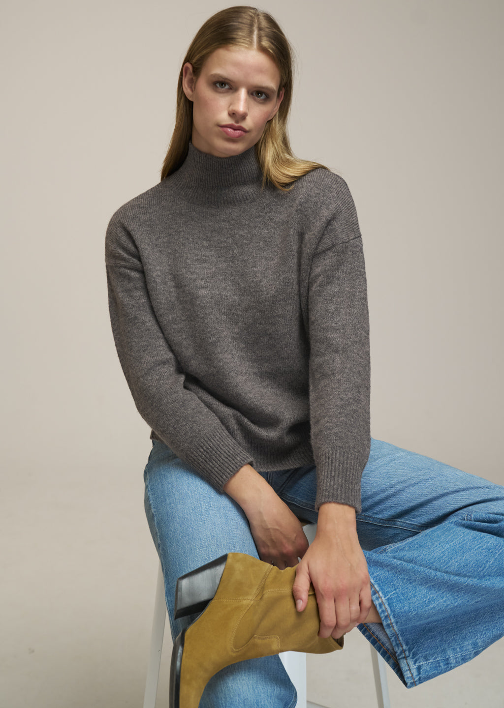 A woman wearing a dark gray turtleneck sweater, blue jeans, and a mustard yellow boot, sitting on a white stool against a plain beige background.