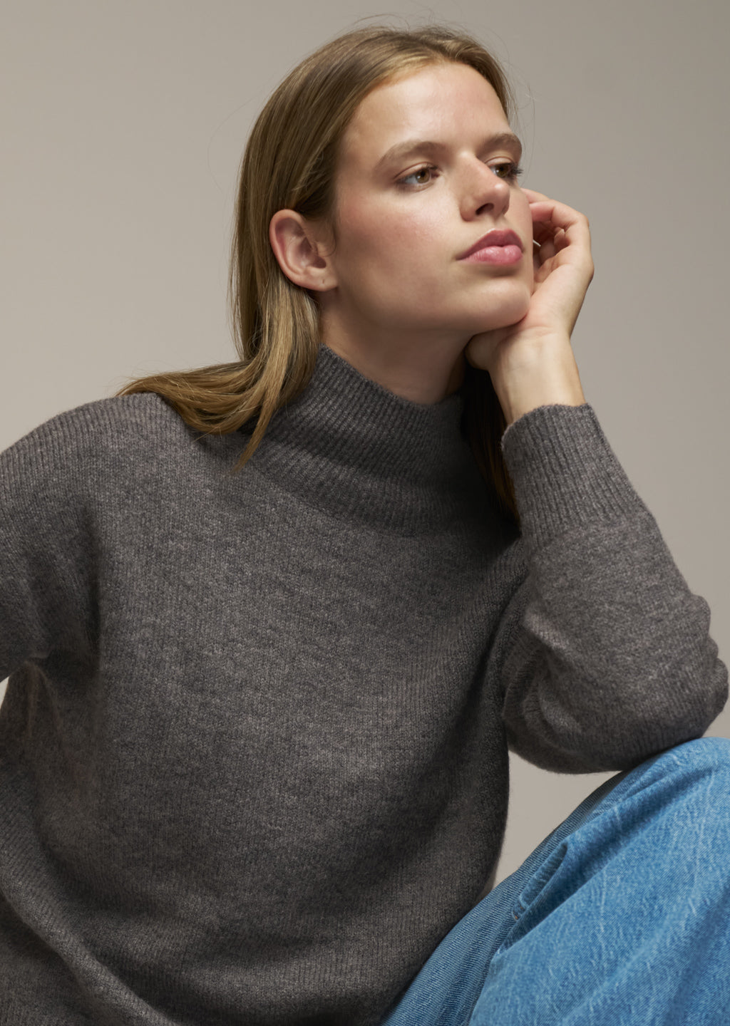 A woman wearing a dark gray ribbed mock neck sweater and blue jeans, posing against a solid neutral gray background.