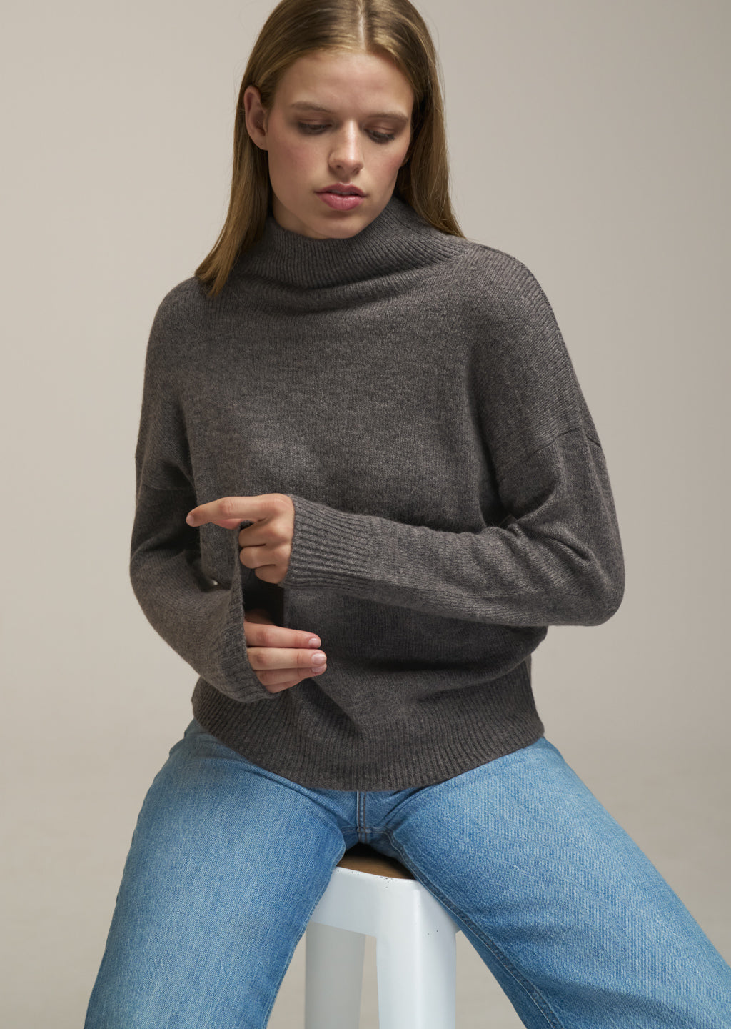 A woman wearing a dark gray ribbed turtleneck sweater and blue jeans, sitting on a white stool against a neutral beige background.