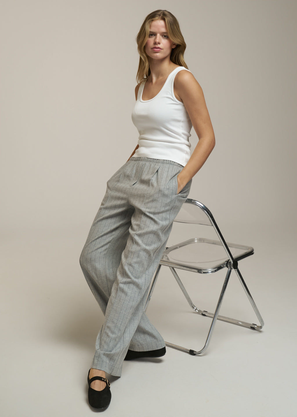 A woman models a pair of light gray wide-leg trousers with a white ribbed tank top and black flats, leaning against a modern metal folding chair against a neutral beige background.