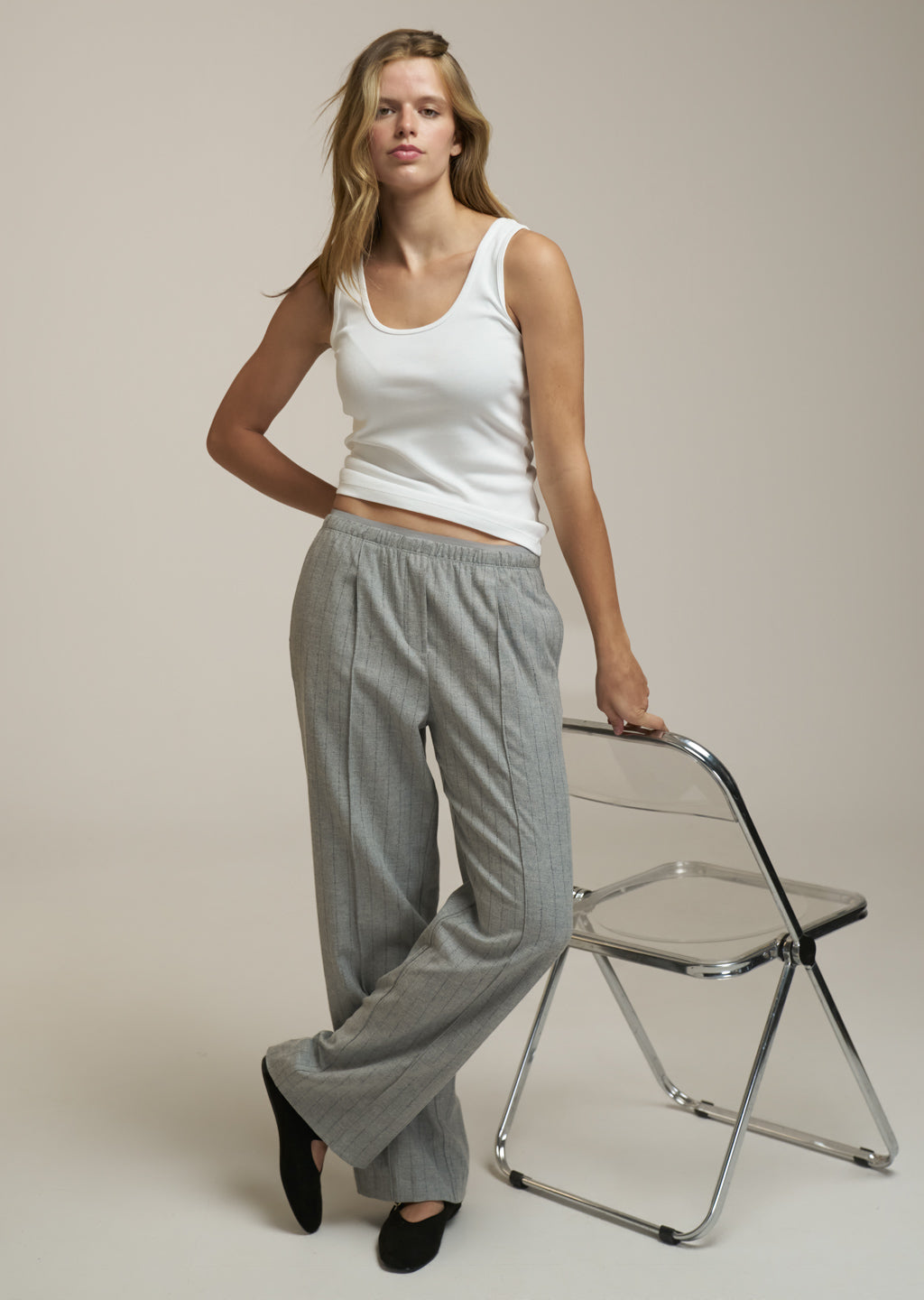 A woman models a pair of light gray wide-leg trousers with an elastic waistband, paired with a white tank top and black flats, standing next to a clear plastic folding chair against a beige studio background.