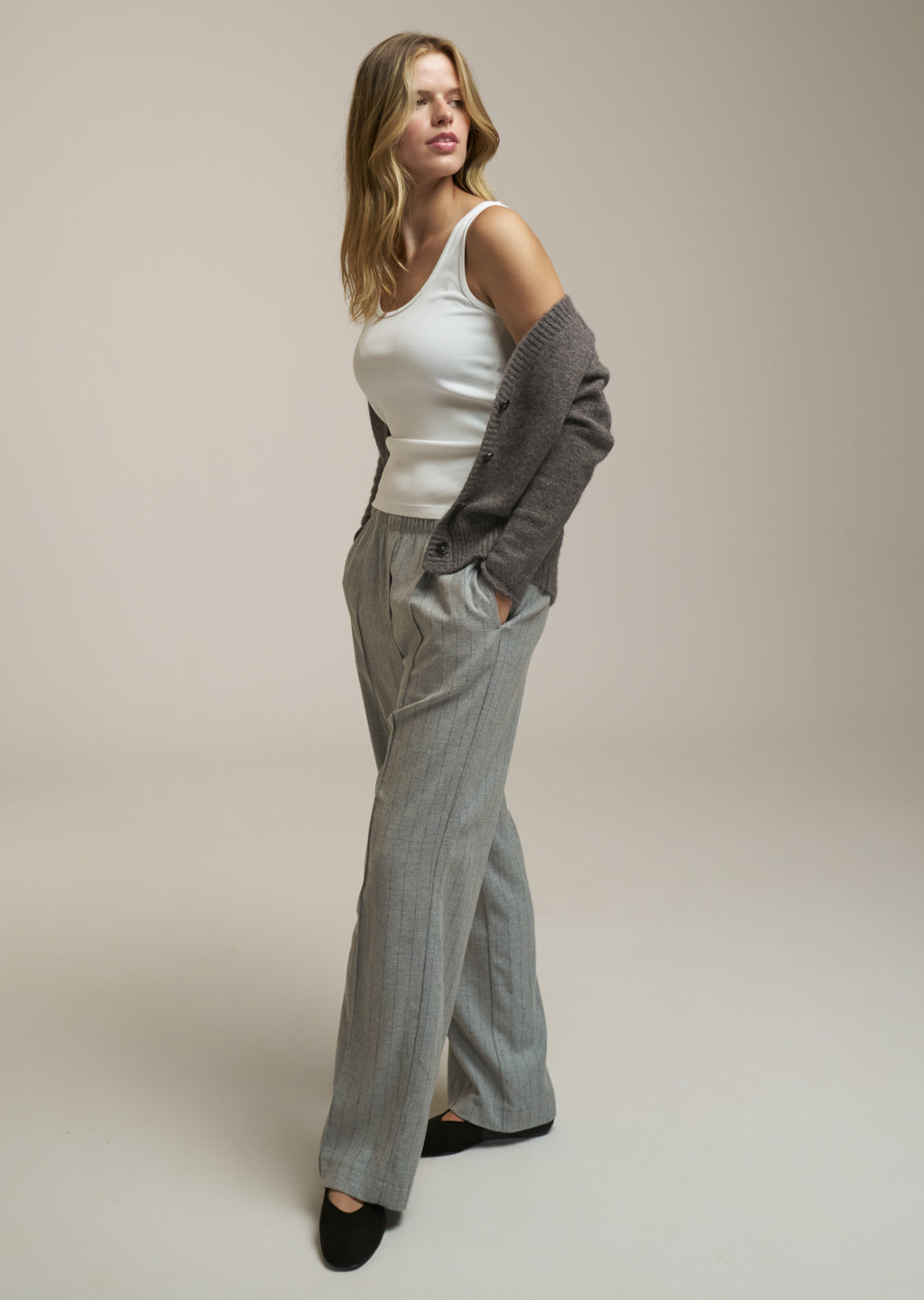A woman modeling a white ribbed tank top, light gray wide-leg trousers, and a dark gray knit cardigan draped off her shoulders, set against a plain beige studio background.