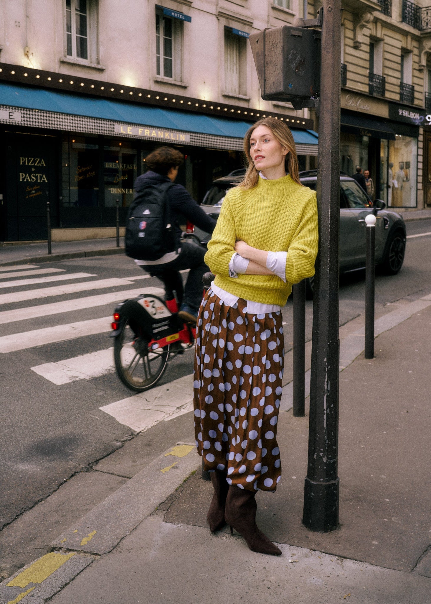 A woman stands on a city street corner wearing a bright yellow ribbed turtleneck sweater and a brown skirt with large white polka dots. A cyclist and parked cars are visible in the background, along with storefronts and a black pole.