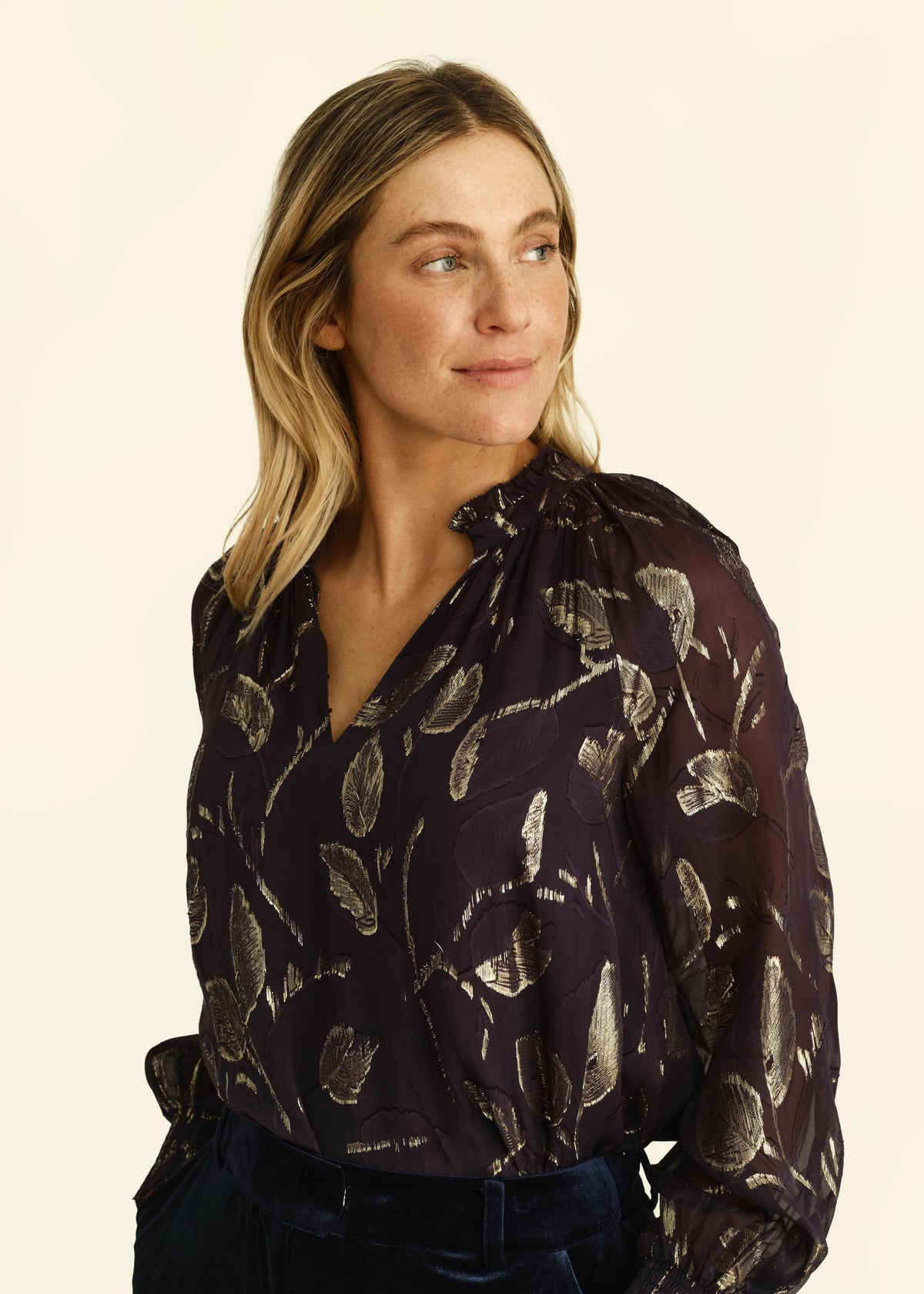 A woman wearing a sheer dark purple blouse with a metallic gold leaf pattern, paired with dark velvet trousers, photographed against a solid cream background.
