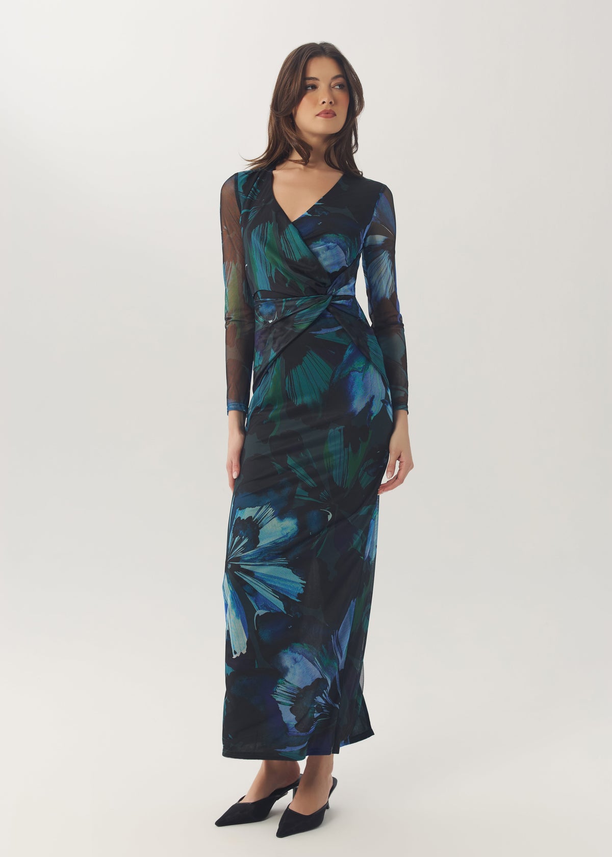 A woman modeling a long-sleeve wrap maxi dress with a dark navy and teal blue floral print, paired with black pointed-toe flats, against a light gray studio background.