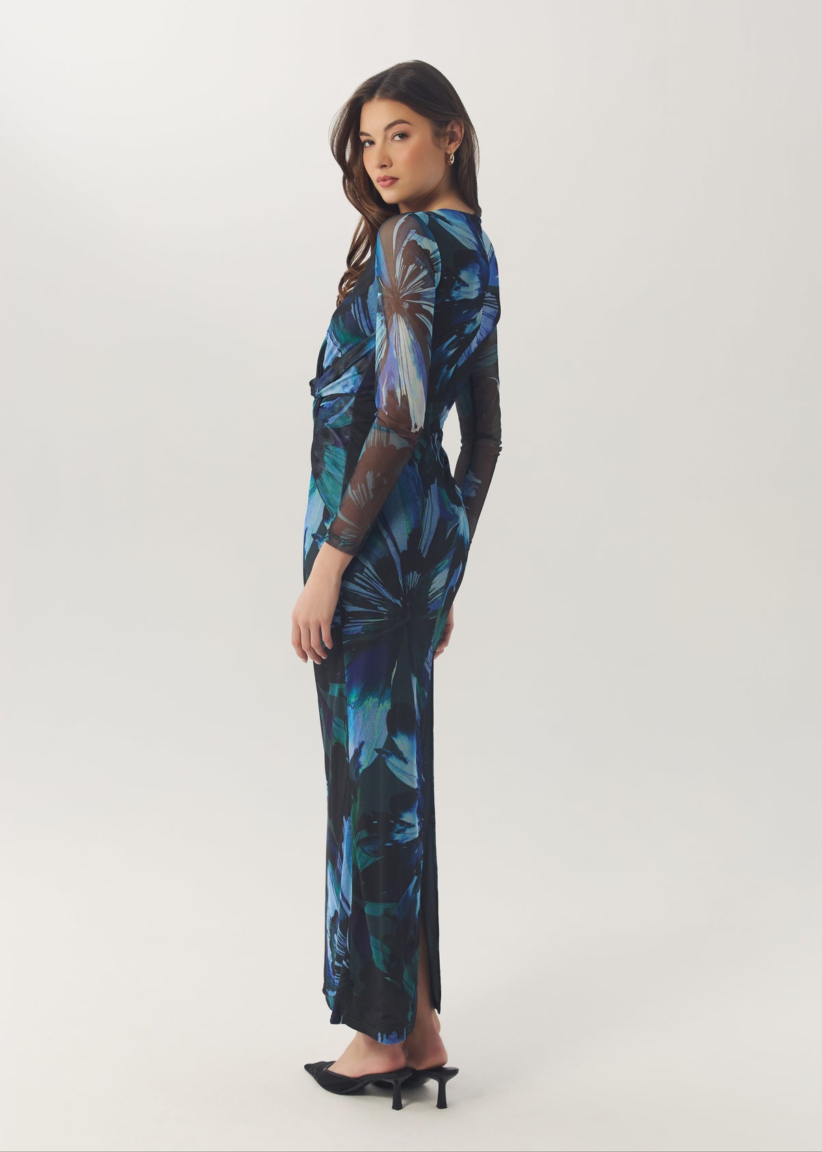 A woman modeling a long-sleeved maxi dress with a blue and black abstract floral print, standing in profile against a light gray studio background.