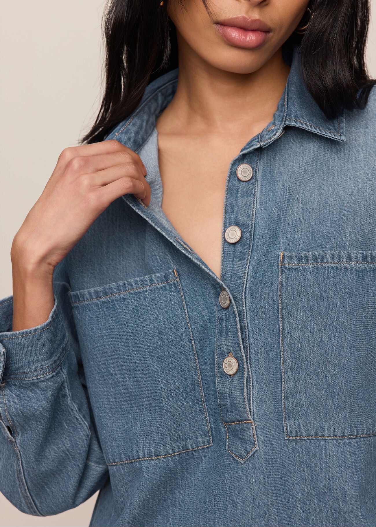 A close-up of a woman wearing a blue denim button-up shirt with two chest pockets and light-colored buttons, set against a plain beige background.