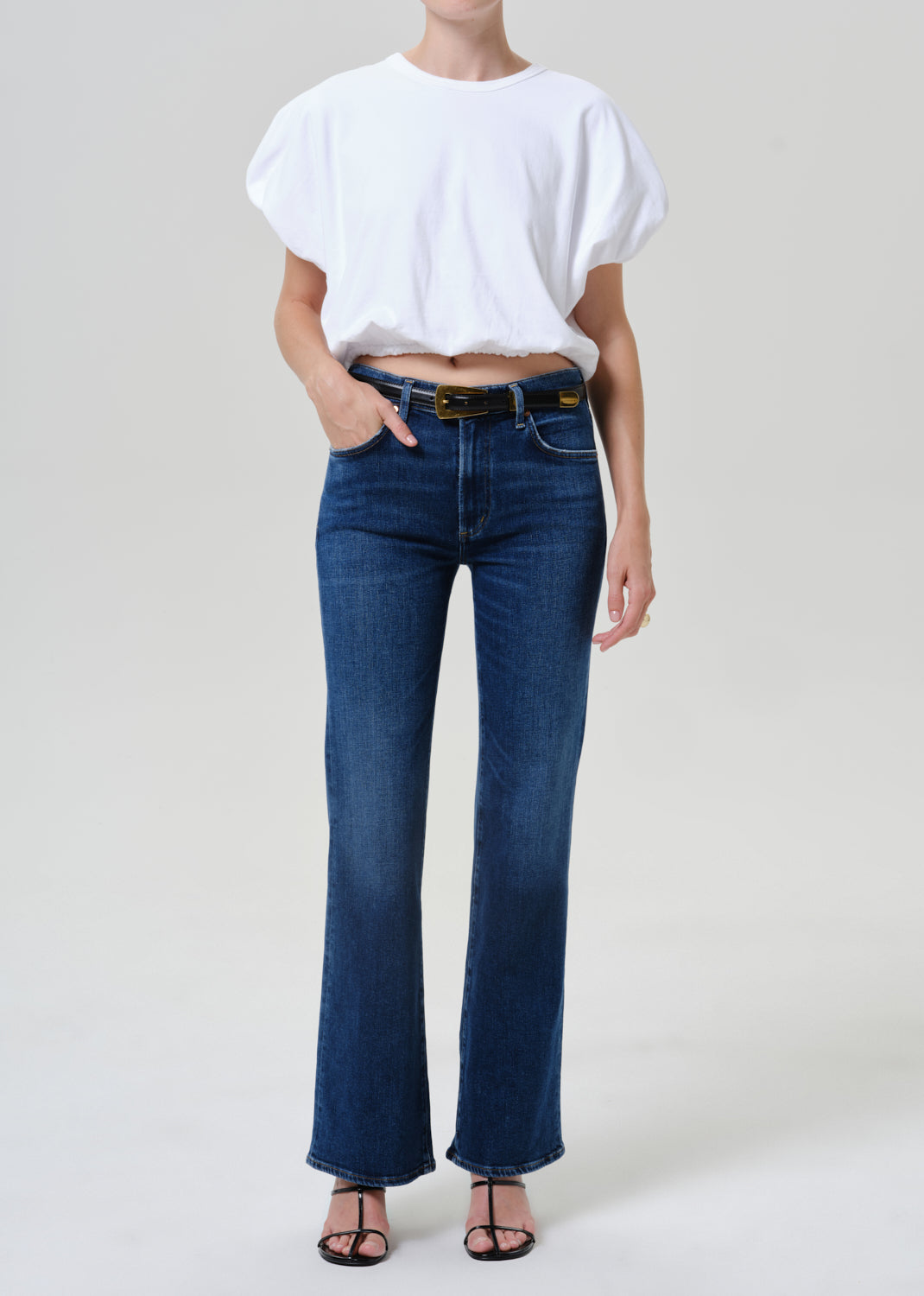 A model wearing dark blue high-waisted flare jeans with a black belt, paired with a white cropped t-shirt and black sandals, photographed against a light gray studio background.