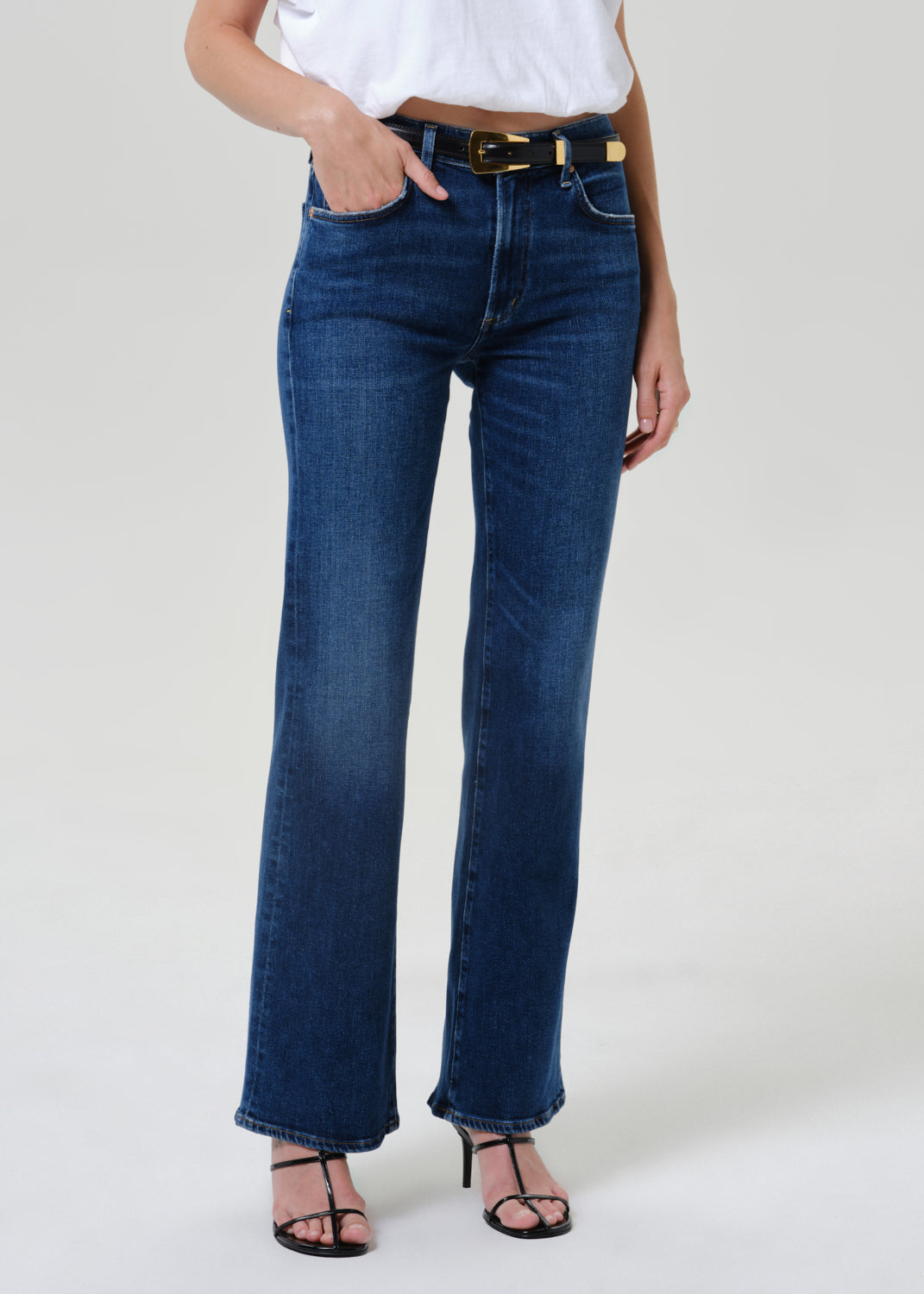 A model wearing a pair of dark blue high-rise flare jeans with a gold-tone belt, paired with a white top and black strappy heels, shown from the waist down on a light gray studio background.