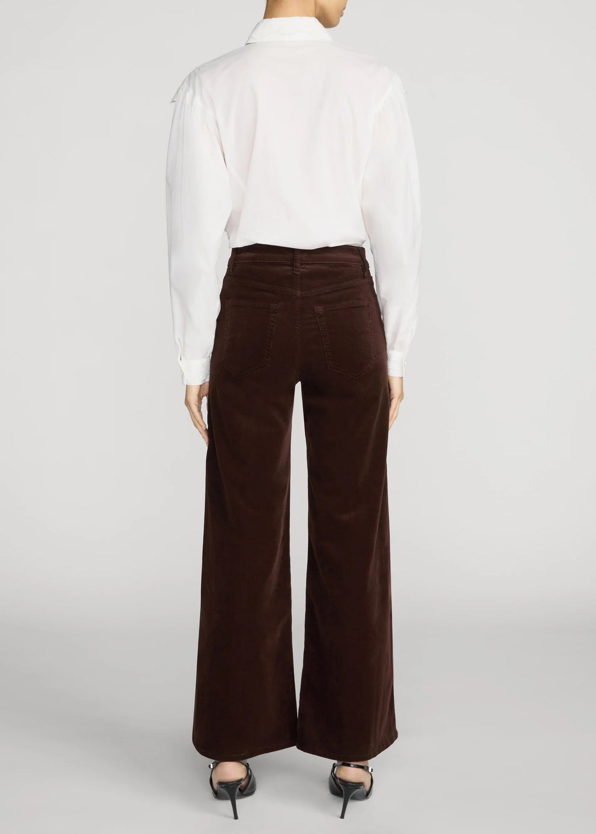 A model shown from the back wearing high-waisted, wide-leg brown corduroy pants, a white long-sleeve button-down shirt, and black pointed-toe heels against a light gray studio background.
