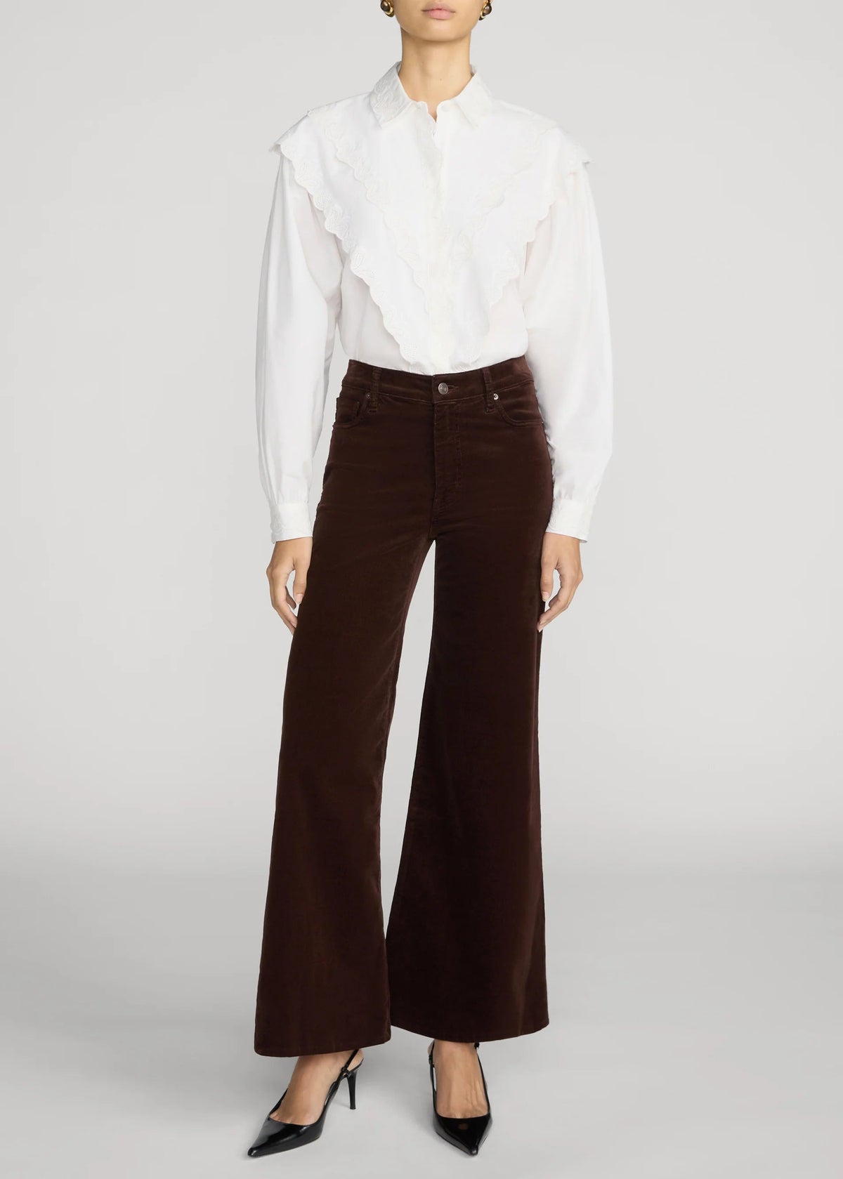 A model wearing a white long-sleeve blouse with ruffle details and dark brown wide-leg corduroy pants, paired with black pointed-toe heels, against a light gray studio background.
