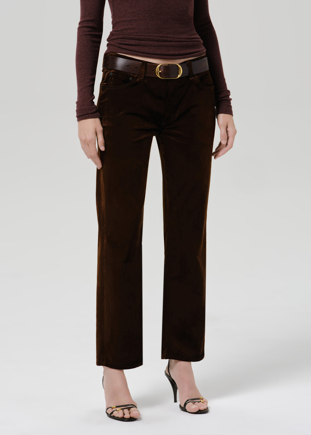 A model wearing dark brown cropped trousers with a matching dark brown belt and a dark brown long-sleeve top, paired with black heeled sandals, against a light gray studio background.