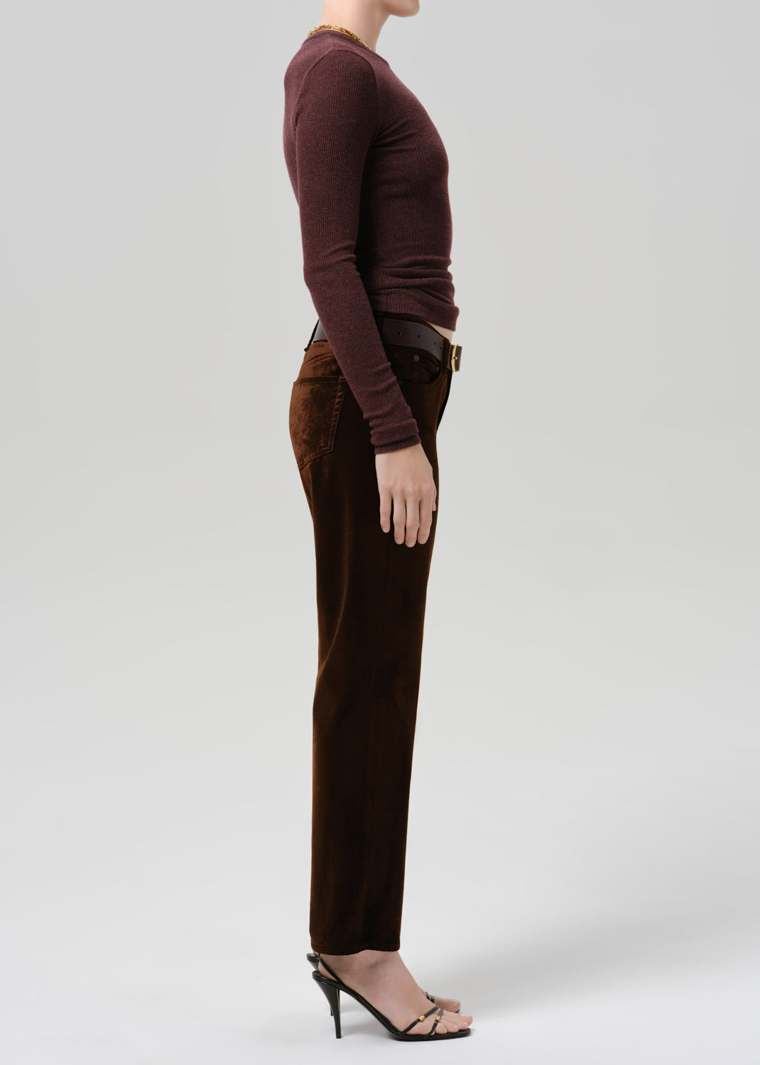 A side-profile view of a model wearing a dark brown corduroy straight-leg pant and a dark plum long-sleeve top, paired with black strappy high-heeled sandals, against a light gray studio background.