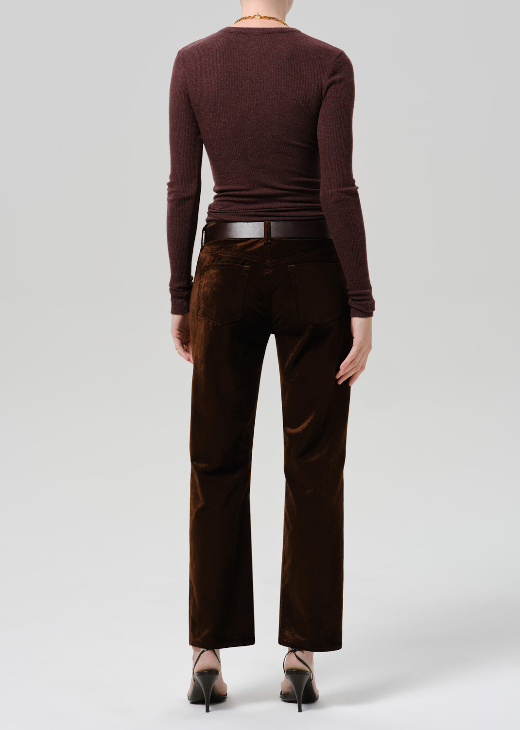 A model shown from the back wearing a dark brown long-sleeve knit top and dark brown wide-leg trousers, styled with a thin black belt and black pointed-toe heels against a light gray studio background.