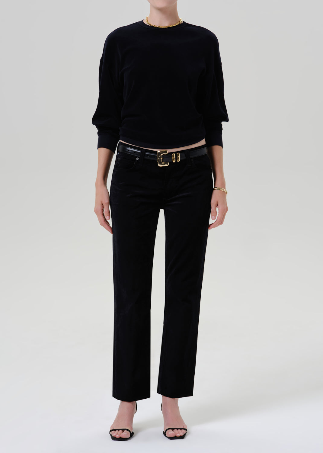 A model wearing a black long-sleeve knit top and black wide-leg trousers with a black belt featuring a gold buckle, photographed against a light gray studio background.