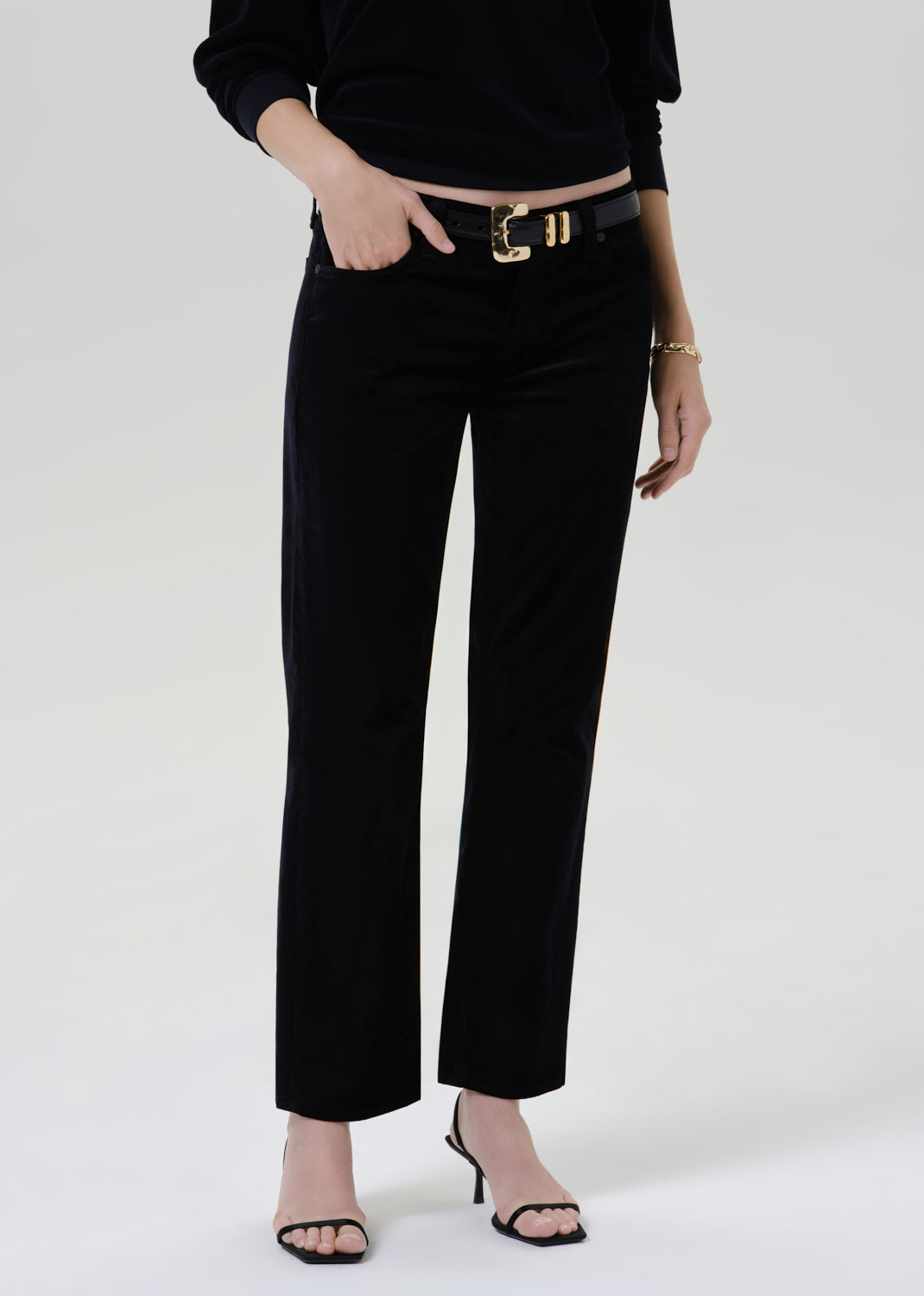 A model wearing black wide-leg cropped trousers with a black belt and gold buckle, styled with a black long-sleeve top and black heeled sandals against a light gray studio background.