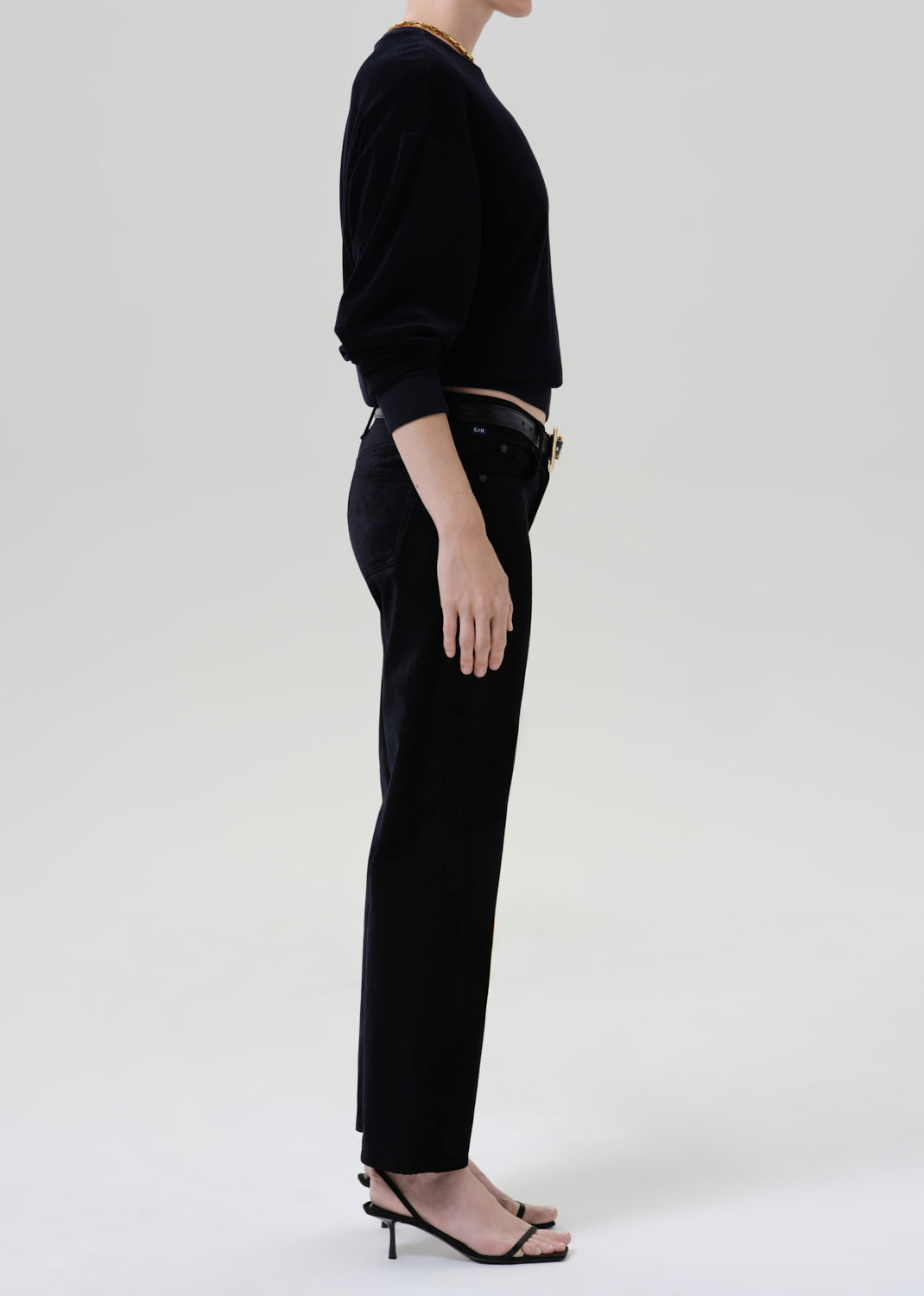 A model shown from the side wearing a black long-sleeve top, black trousers, and black strappy high-heeled sandals against a plain light gray studio background.