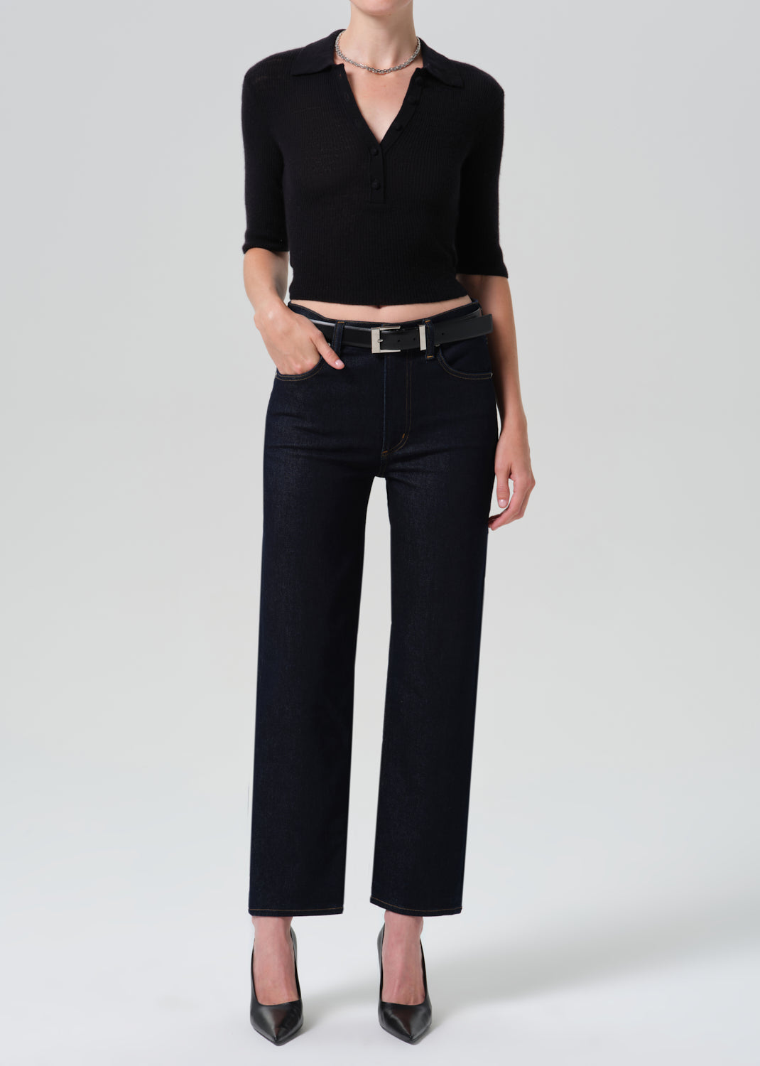 A model wearing a black three-quarter sleeve collared knit top and black wide-leg cropped jeans with a black belt, paired with black pointed-toe heels, against a light gray studio background.