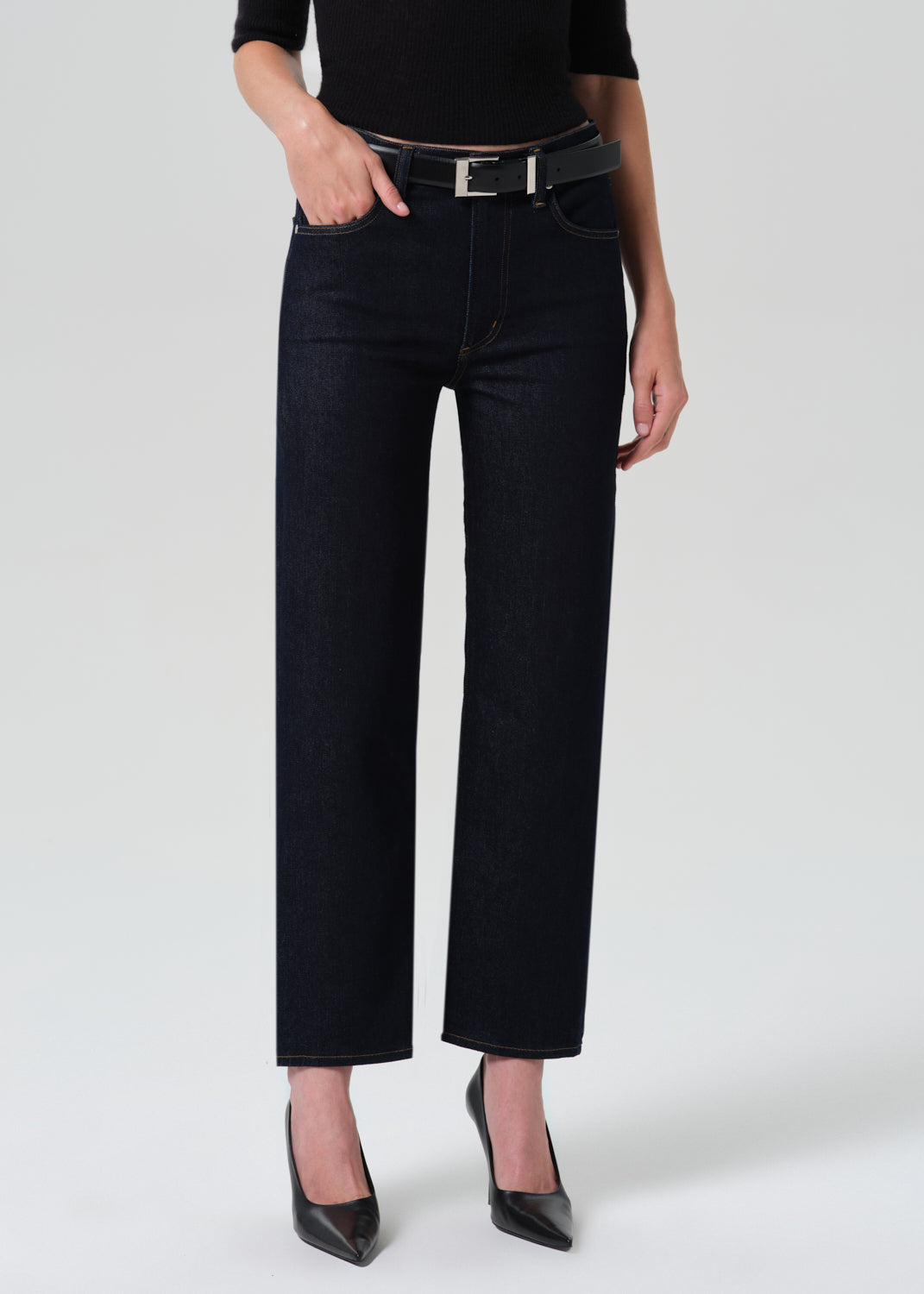 A model wearing black wide-leg cropped jeans with a black belt, paired with a black top and black pointed-toe heels, photographed against a light gray studio background.