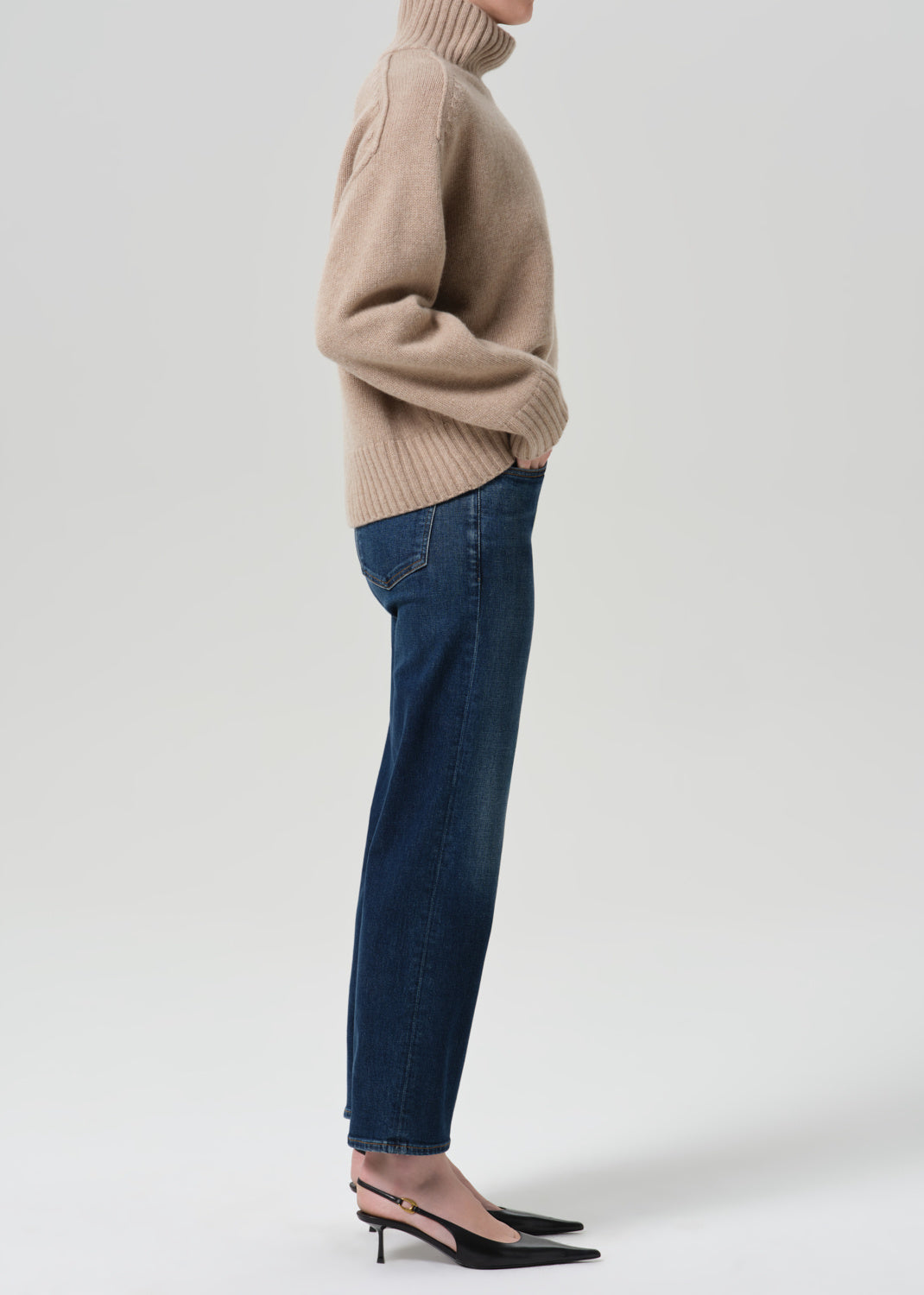 A side-profile studio shot of a model wearing a beige ribbed turtleneck sweater, dark blue straight-leg jeans, and black slingback heels against a light gray background.