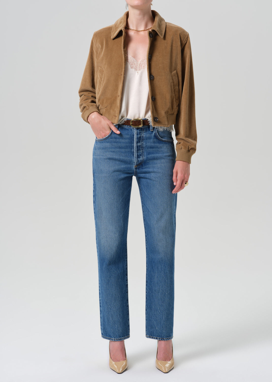 Front view of a model wearing blue straight-leg jeans with a raw, frayed hem, paired with a brown suede jacket and beige heels, against a light gray studio background.