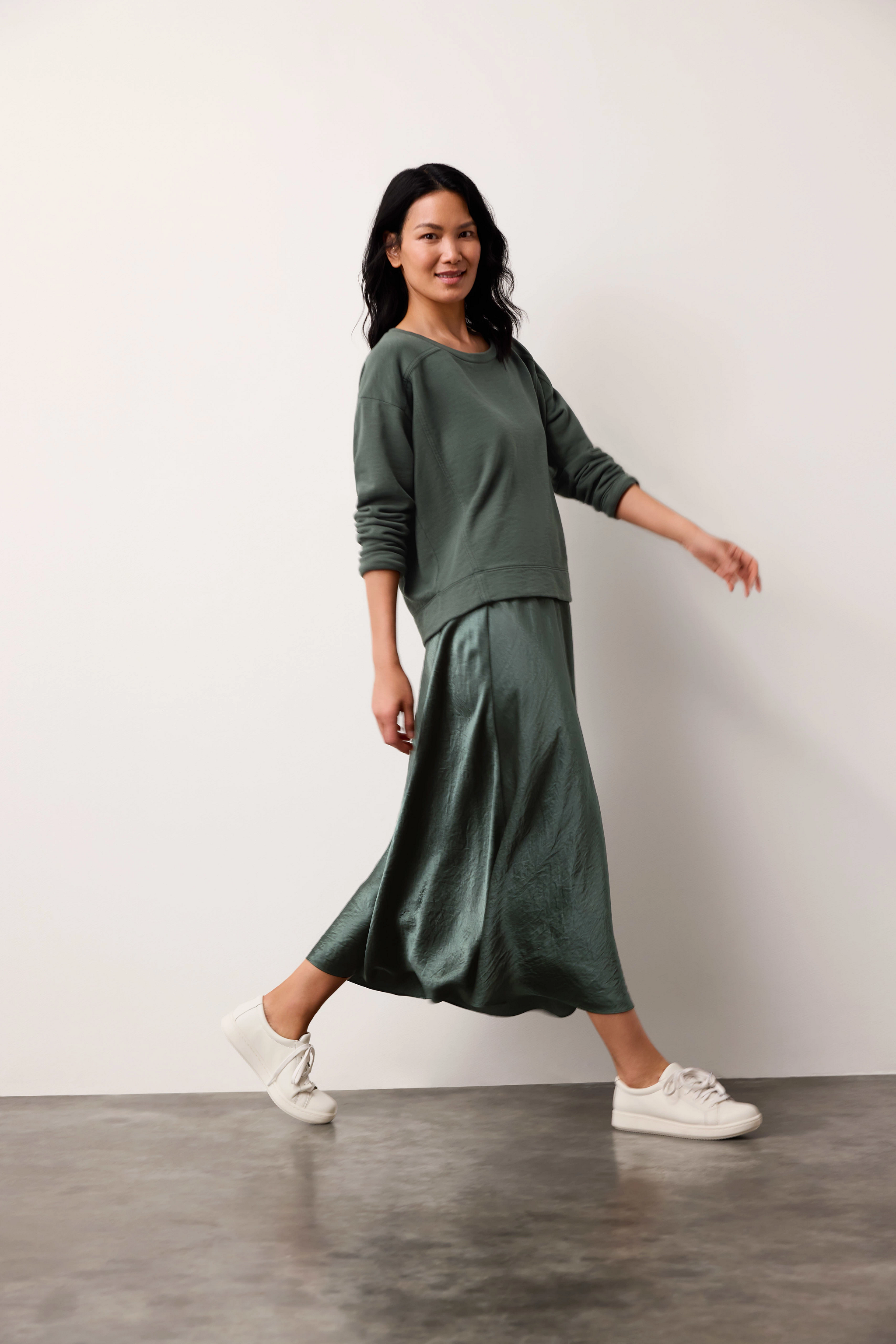 Woman wearing an Eileen Fisher skirt in  green in the color Everglade with white sneakers on a plain background