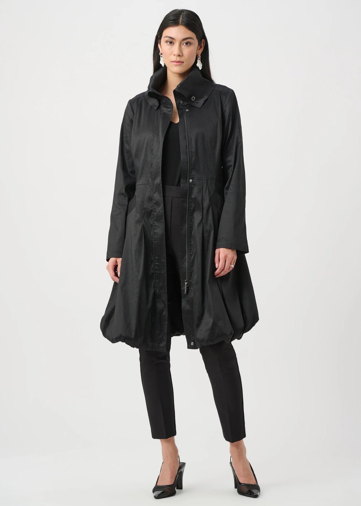 A model stands against a light gray studio background wearing a long black coat over a black outfit with black cropped pants and black heels.