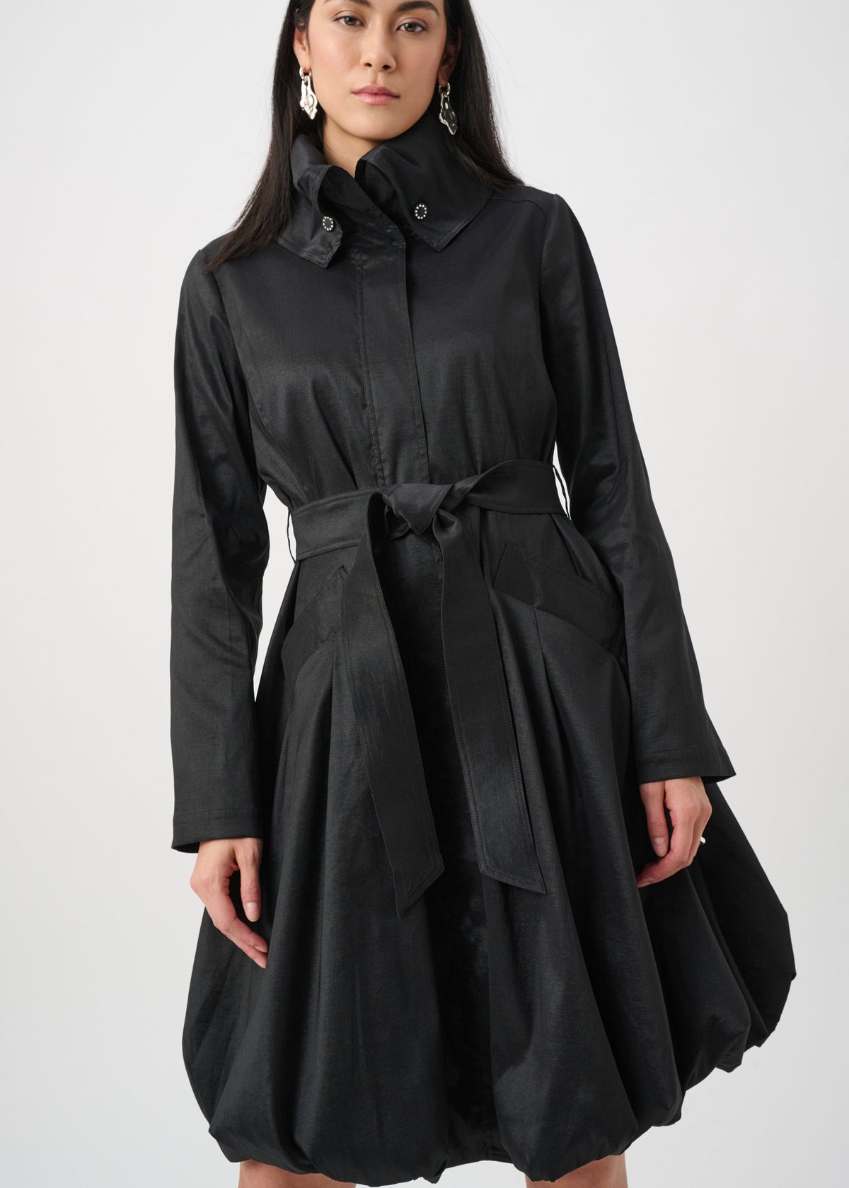 A woman modeling a black long-sleeve collared dress with a matching waist tie and flared skirt against a plain light gray studio background.