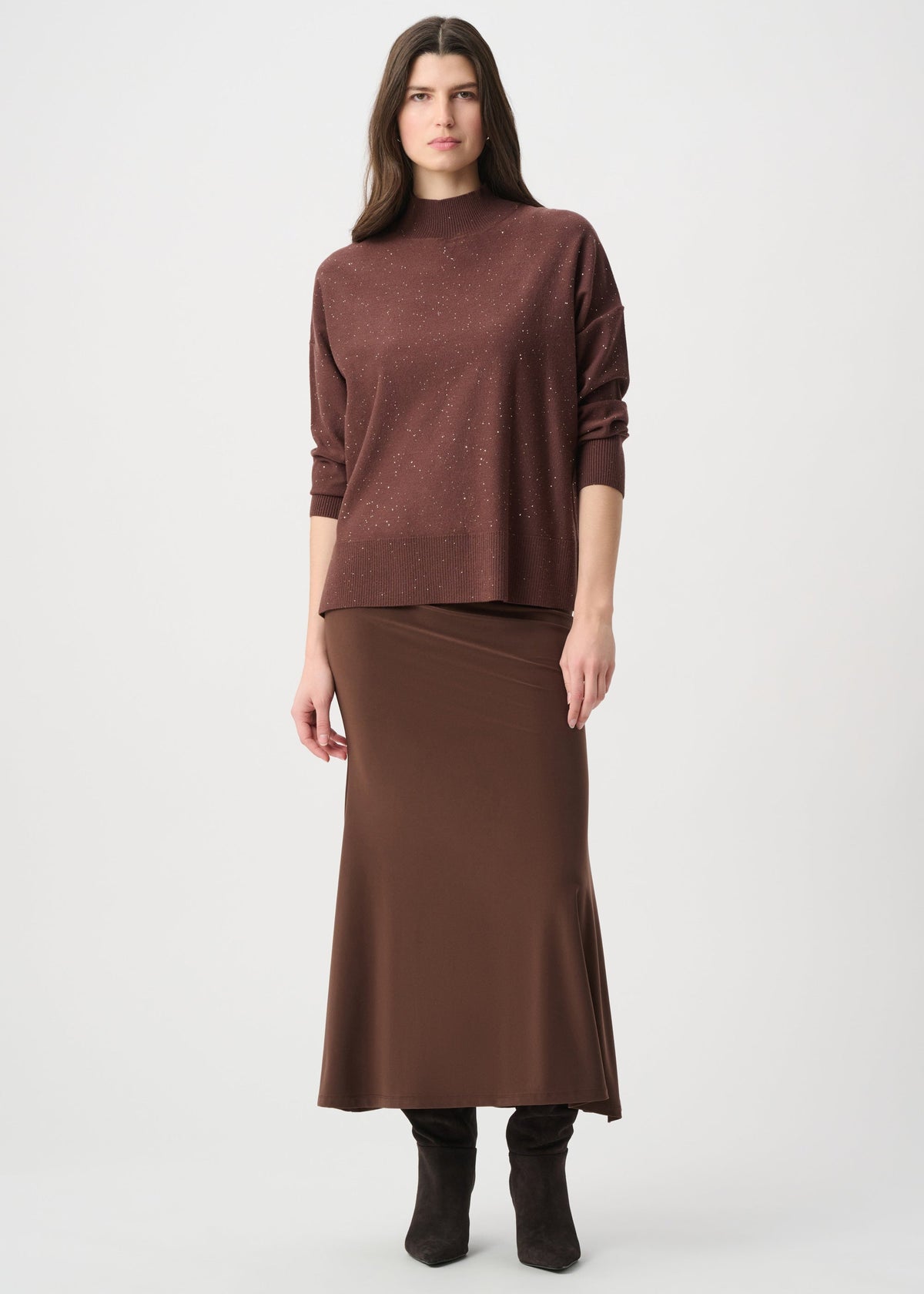 A model stands against a light gray studio background wearing a dark brown long-sleeve knit top and a matching brown midi skirt, paired with black ankle boots.