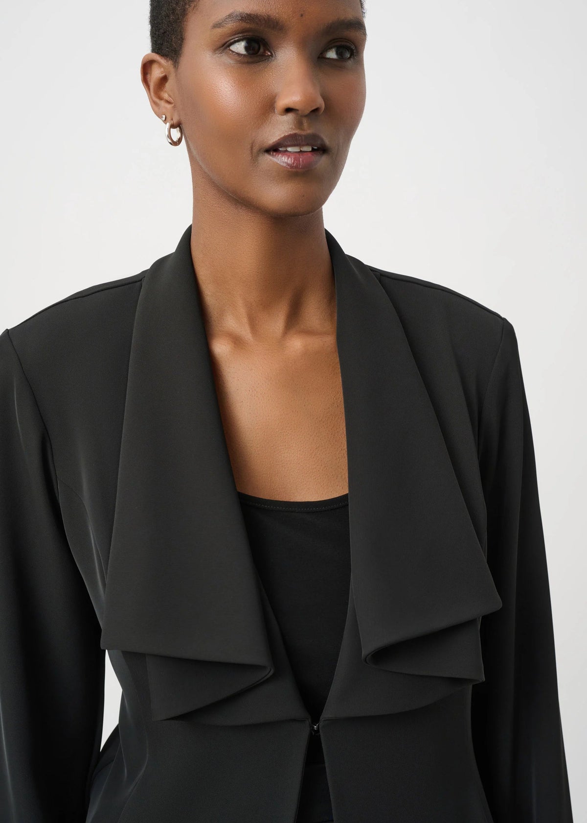 A close-up of a model wearing a black draped wrap-front blazer over a black top, set against a plain light gray studio background.