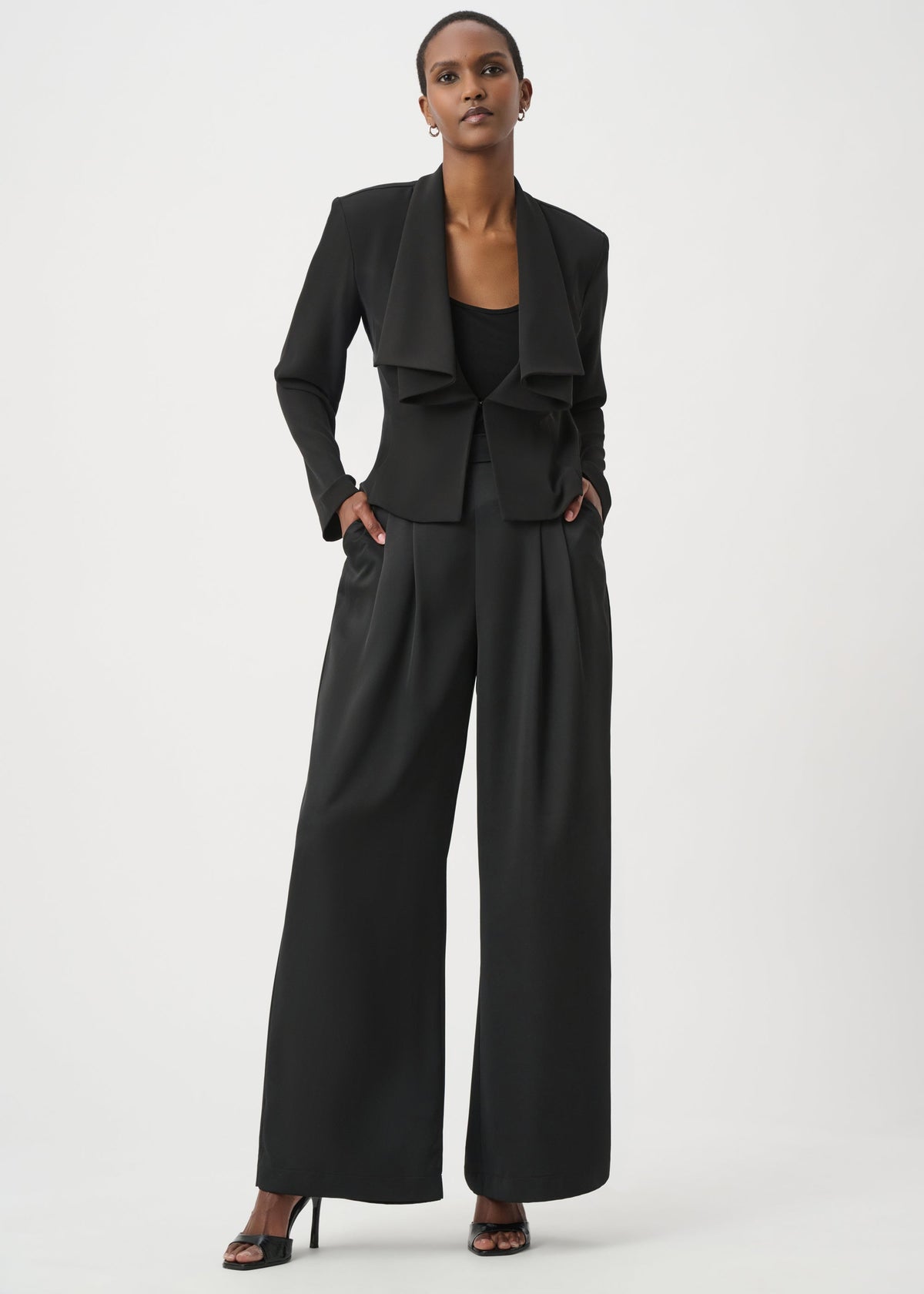A model wearing a black ruffled blazer and matching wide-leg trousers, styled with black heels against a plain light gray studio background.