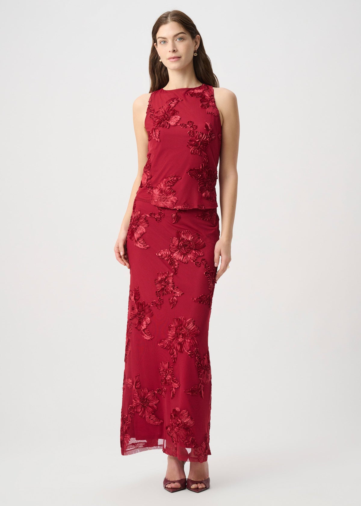 A woman modeling a sleeveless, high-neck, floor-length red dress with tonal floral lace and sequin applique, photographed against a plain light gray studio background.