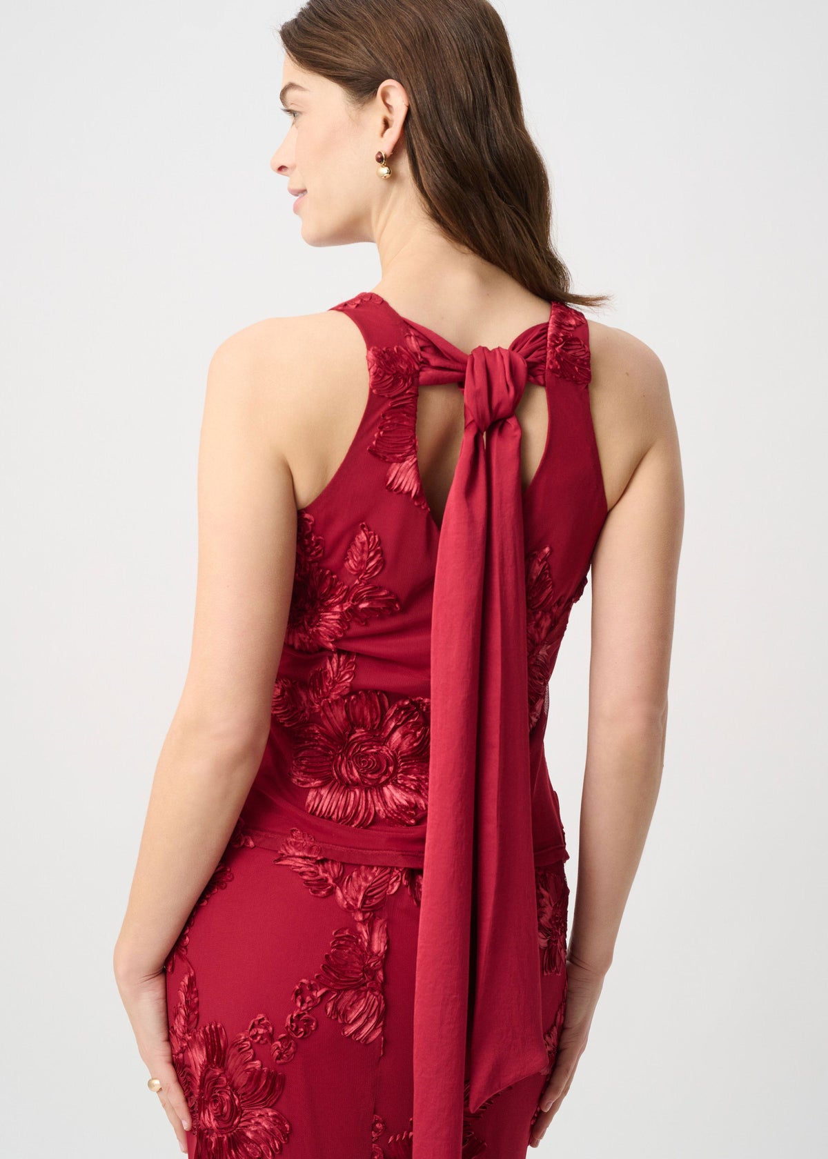 A woman shown from the back wearing a sleeveless deep red dress with floral embroidery and a long tied bow detail at the neckline, photographed against a light gray studio background.