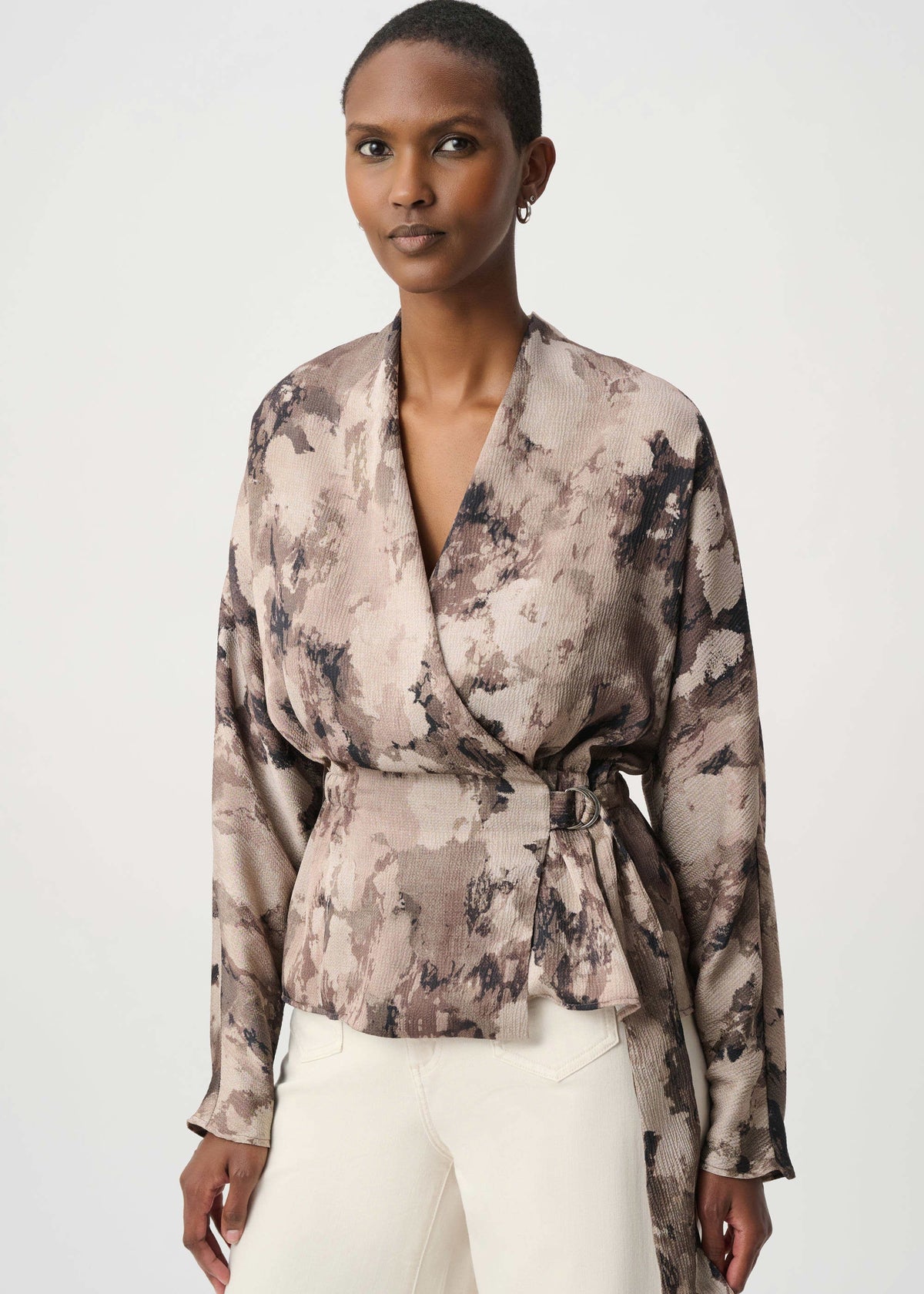 A woman modeling a long-sleeve wrap-style blouse with an abstract watercolor print in beige, taupe, and charcoal gray, paired with white pants against a light gray studio background.