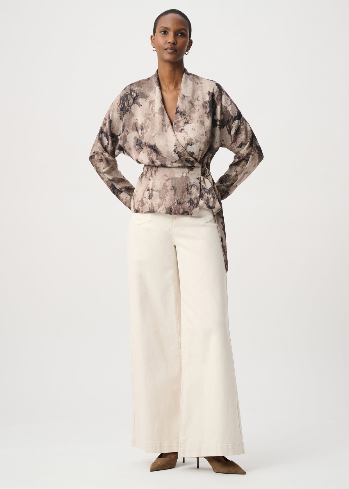 A model stands against a light gray studio background wearing a long-sleeve belted wrap top with a brown and beige abstract print, paired with wide-leg cream trousers and brown pointed-toe heels.