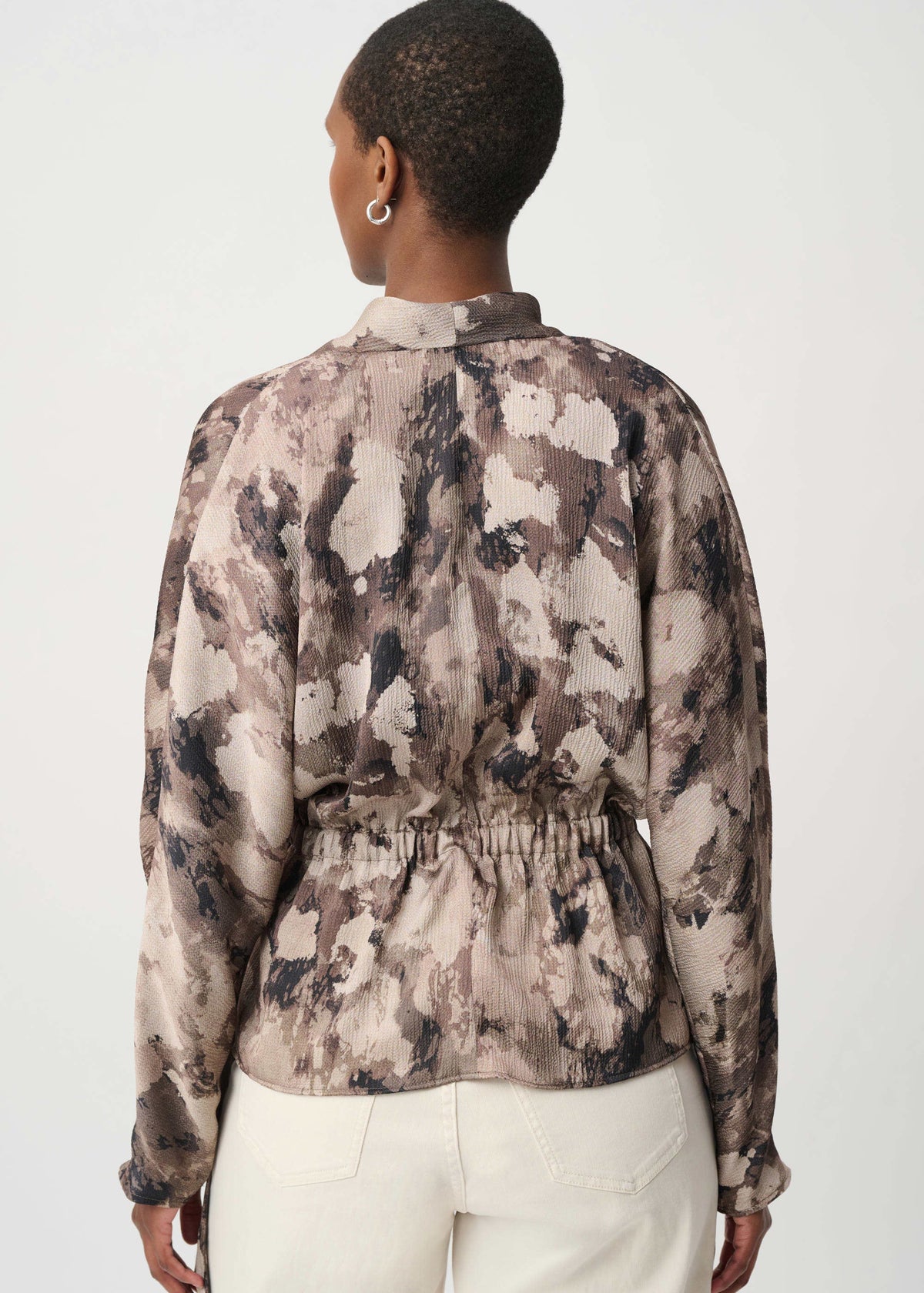A woman shown from the back wearing a long-sleeve collared top with an abstract watercolor-style print in beige, taupe, brown, and black, paired with white pants, against a plain light gray studio background.