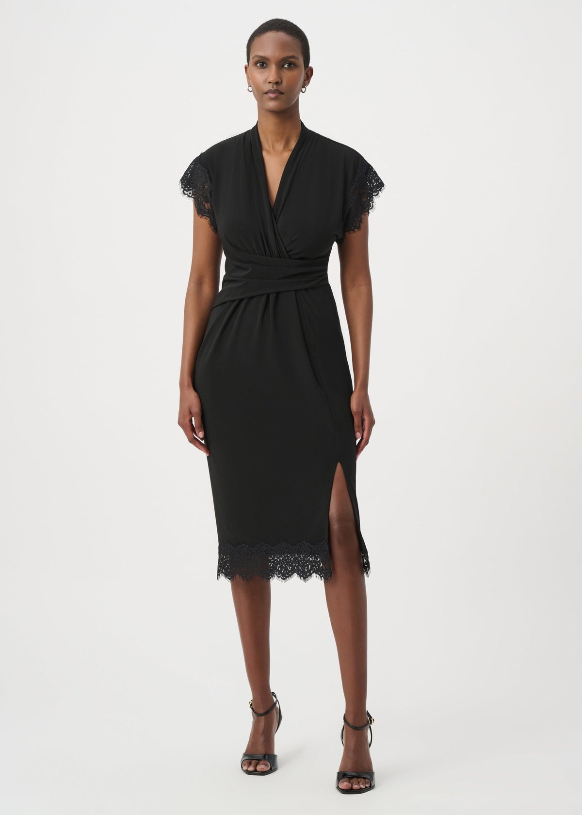 A model wearing a black midi wrap dress with lace trim on the sleeves and hem, paired with black strappy high-heeled sandals, against a plain light gray studio background.