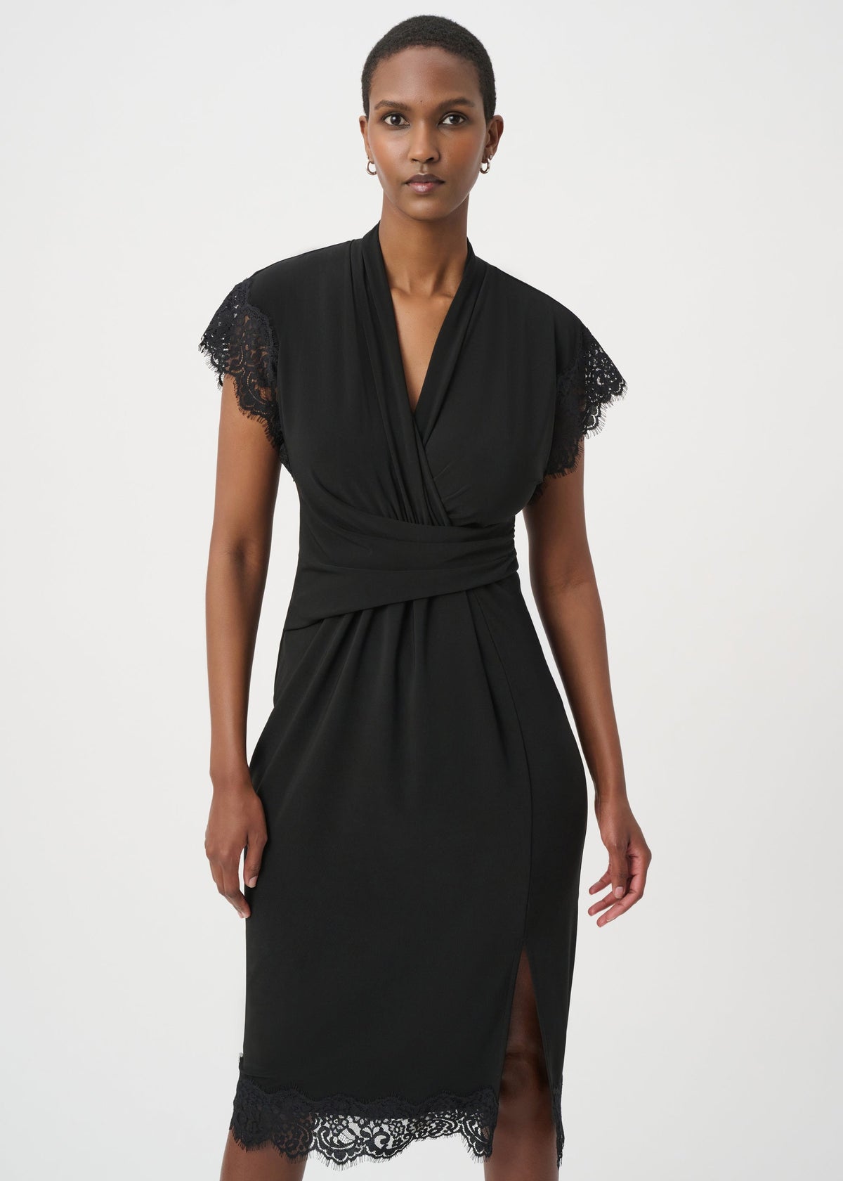 A woman modeling a black wrap-style midi dress with short cap sleeves, lace trim at the sleeve cuffs and hem, and a side slit, photographed against a plain light gray studio background.