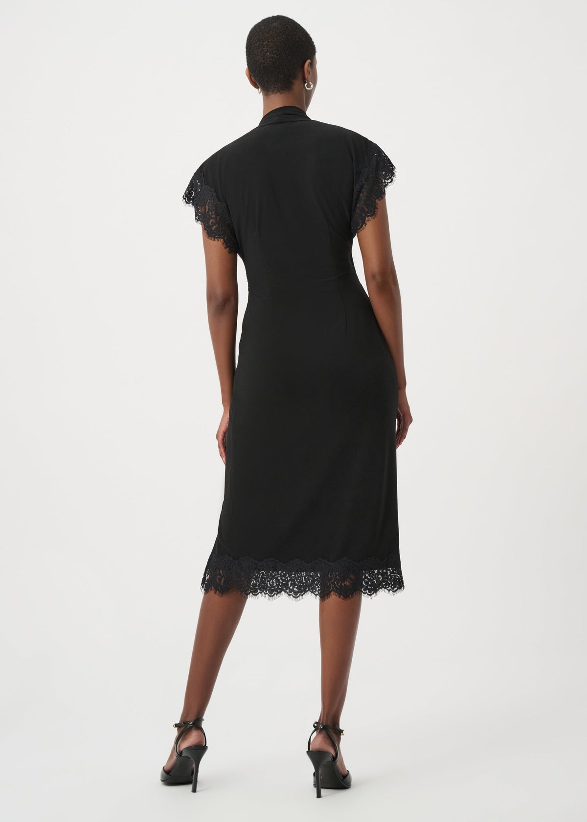 A woman viewed from the back wearing a black midi dress with short cap sleeves and lace trim at the shoulders and hem, paired with black high heels against a plain light gray studio background.