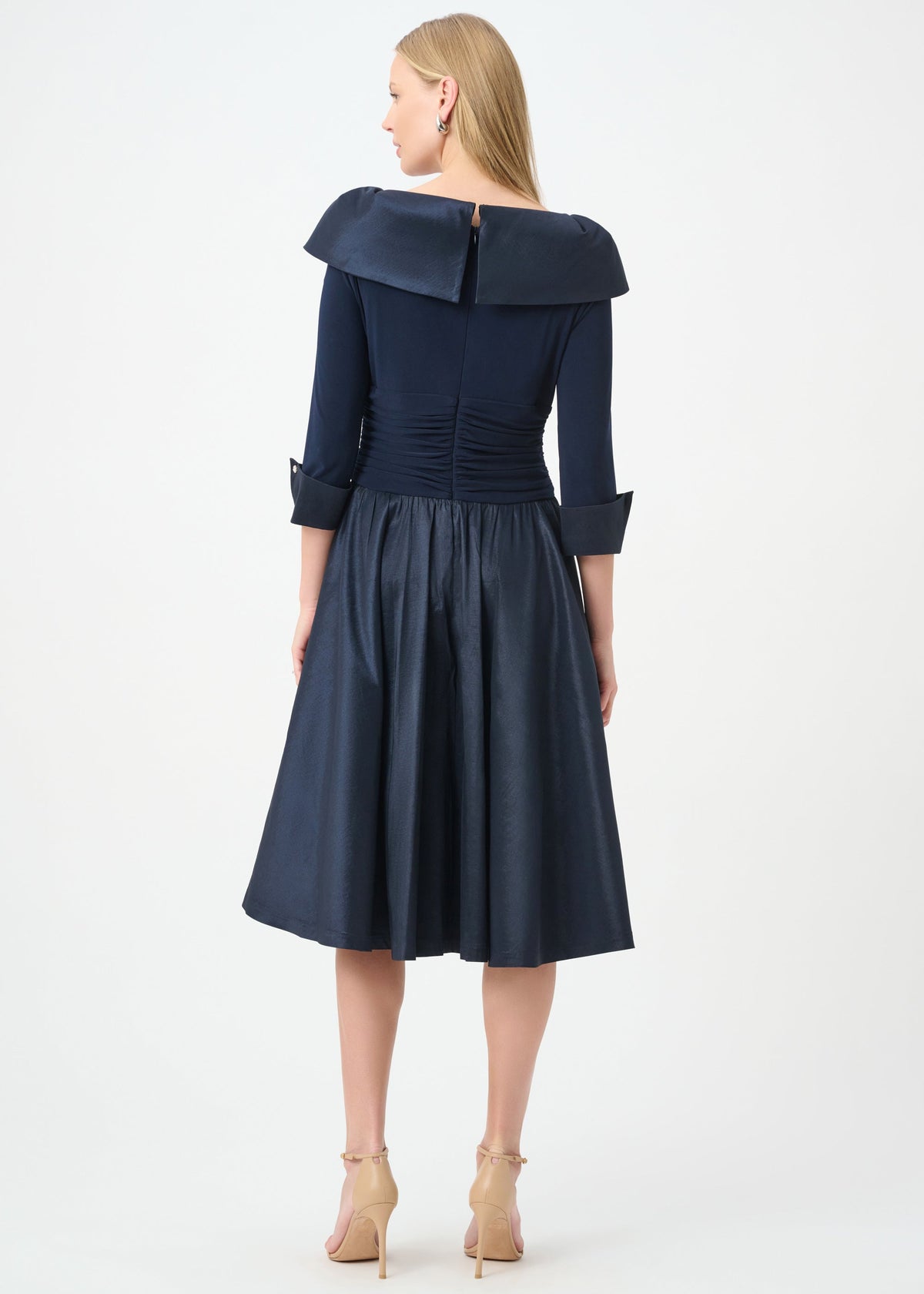 A woman shown from the back wearing a navy blue off-the-shoulder midi dress with three-quarter sleeves, a gathered waist, and a flared skirt, paired with beige high-heeled sandals against a light gray studio background.