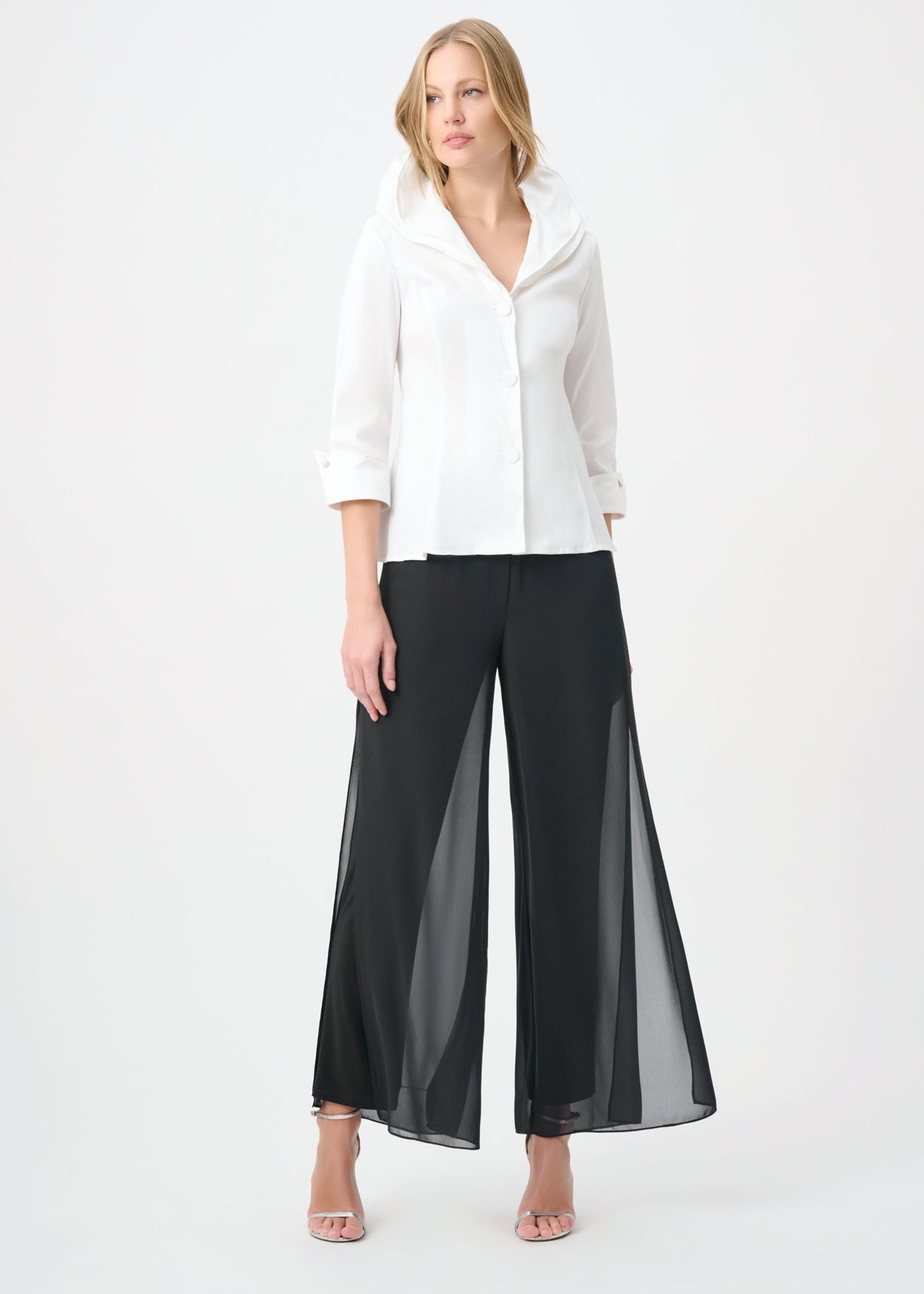 A woman modeling a white button-up blouse with three-quarter sleeves and black wide-leg pants featuring sheer side panels, standing against a plain white studio background.