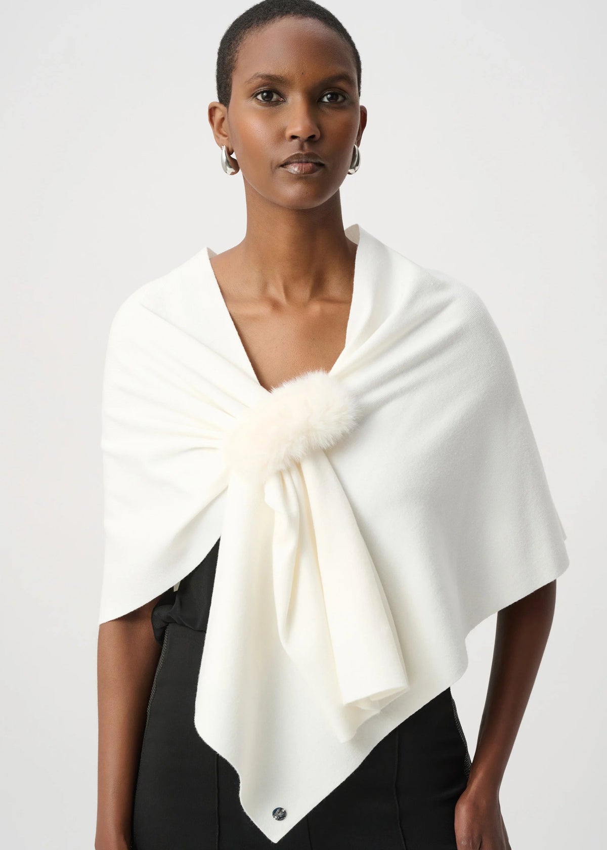 A model wearing a cream-colored wrap shawl with a fluffy pom-pom detail at the center, styled over a black top against a light gray studio background.